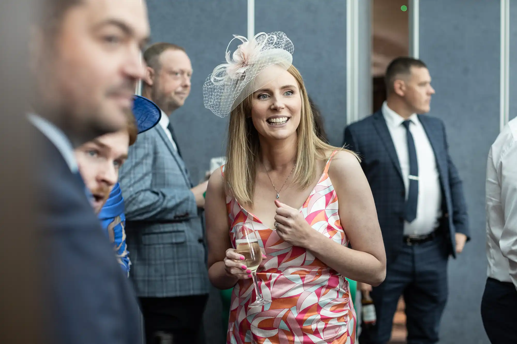 A woman in a colorful dress and fascinator holds a drink and smiles while standing among a group of people dressed in formal attire.