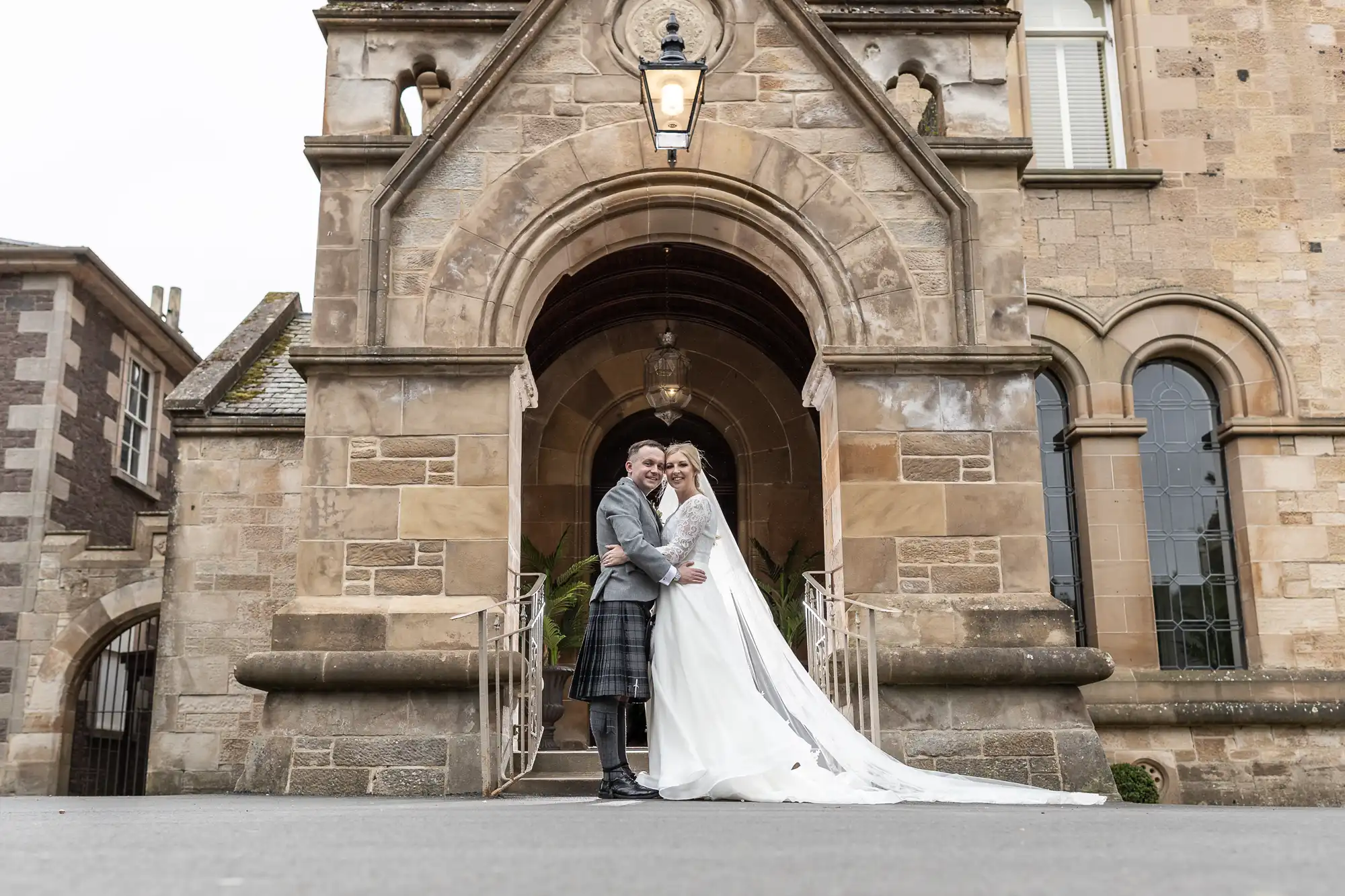 A couple in wedding attire stands on the steps in front of a stone building with an arched entrance. The groom is in a kilt, and the bride is in a long white gown with a train.