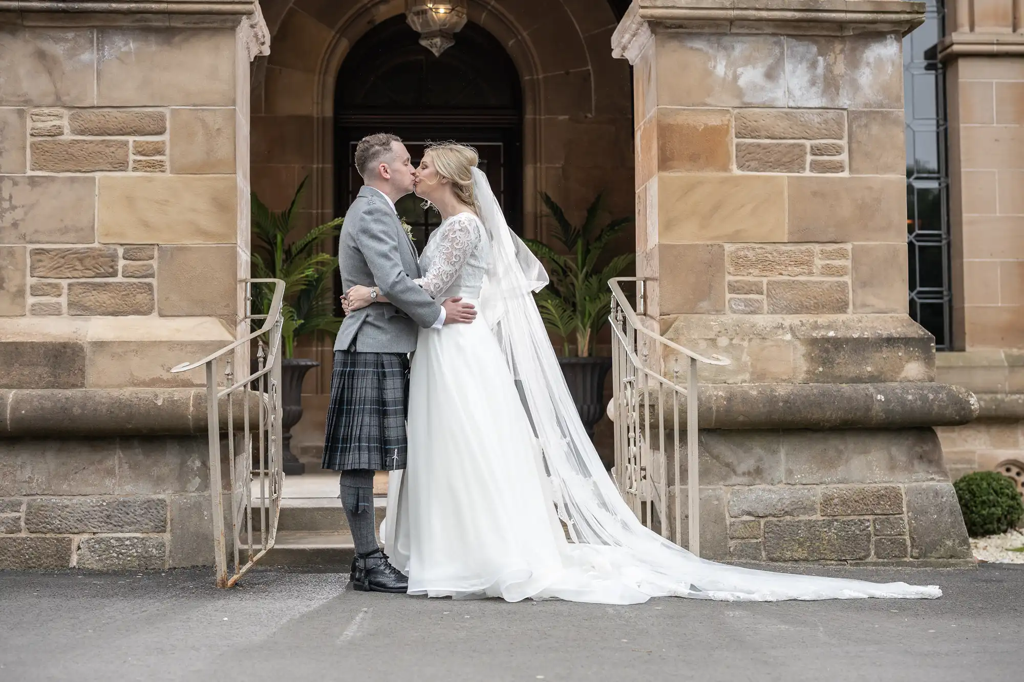 A bride and groom share a kiss in front of a stone building. The bride wears a white dress and veil, and the groom wears a gray jacket and kilt.