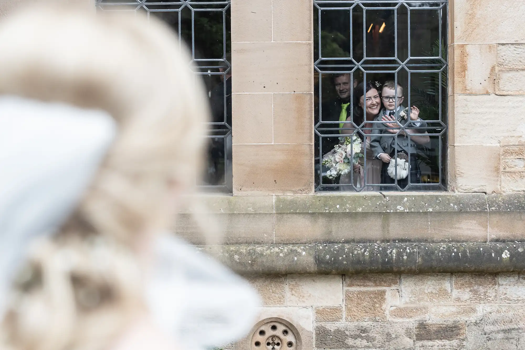 A blurry bride faces a window where three people holding a bouquet and baby wave back from inside a stone building.