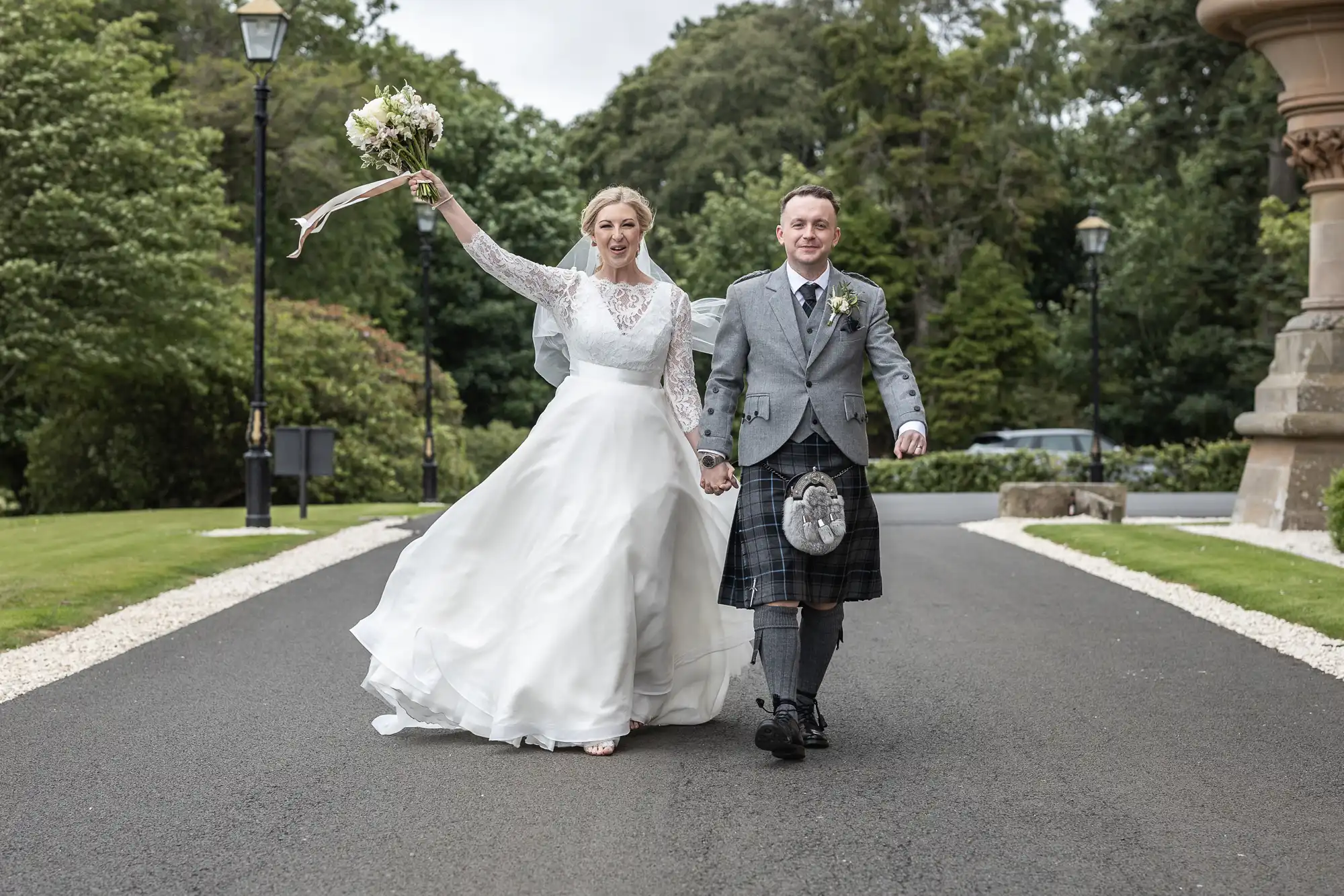 A bride and groom walk hand in hand on a paved path. The bride wears a white gown and holds a bouquet. The groom is dressed in a grey jacket and kilt. Trees and a stone structure are in the background.