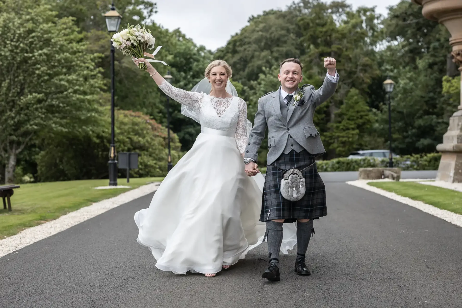 Cornhill Castle wedding photos: A bride and groom walk hand-in-hand outdoors. The bride wears a white dress and holds a bouquet in the air. The groom wears a kilt and gray jacket, raising his fist in celebration. They are smiling.