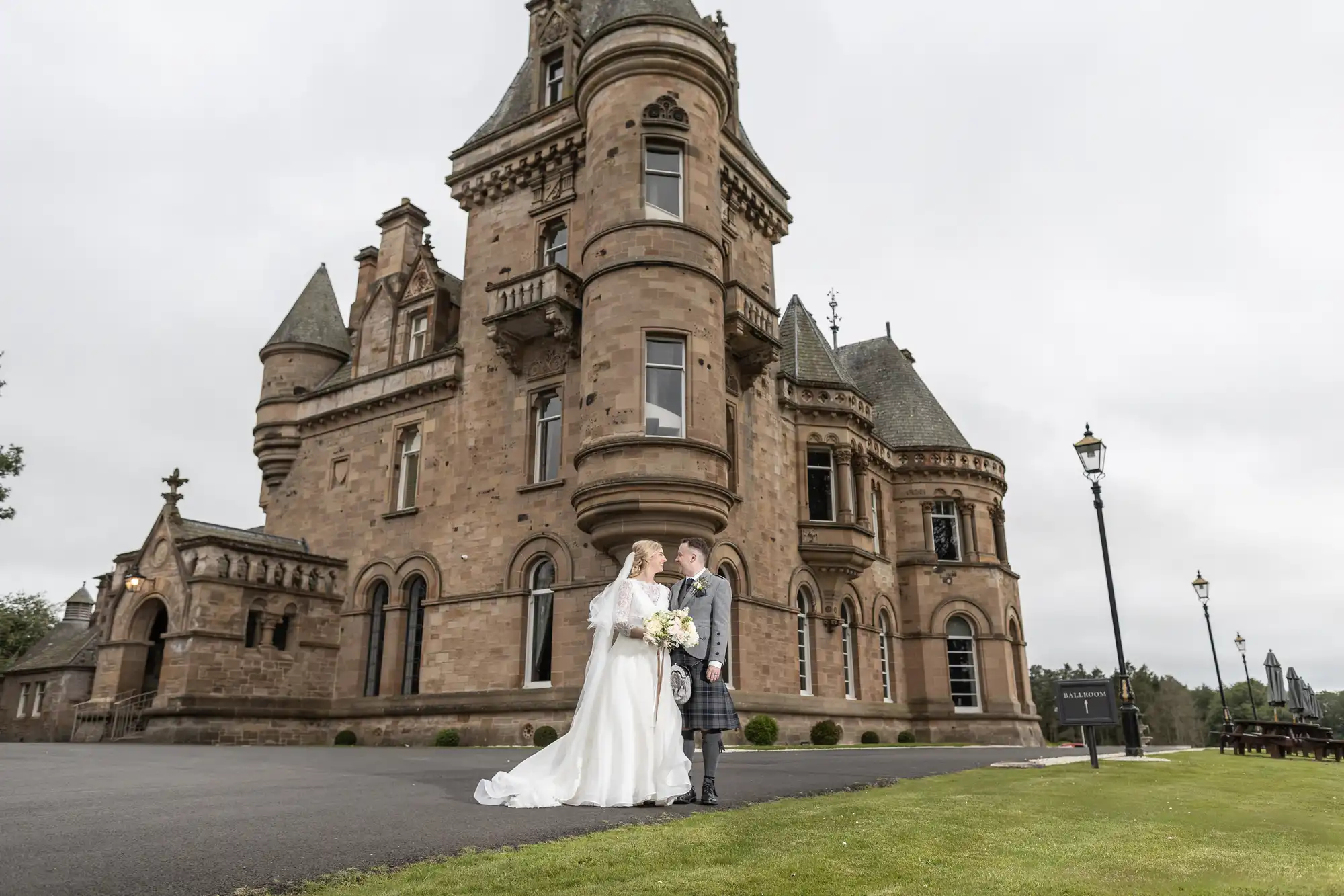 A couple dressed in wedding attire stand hand in hand in front of a large, historic castle.