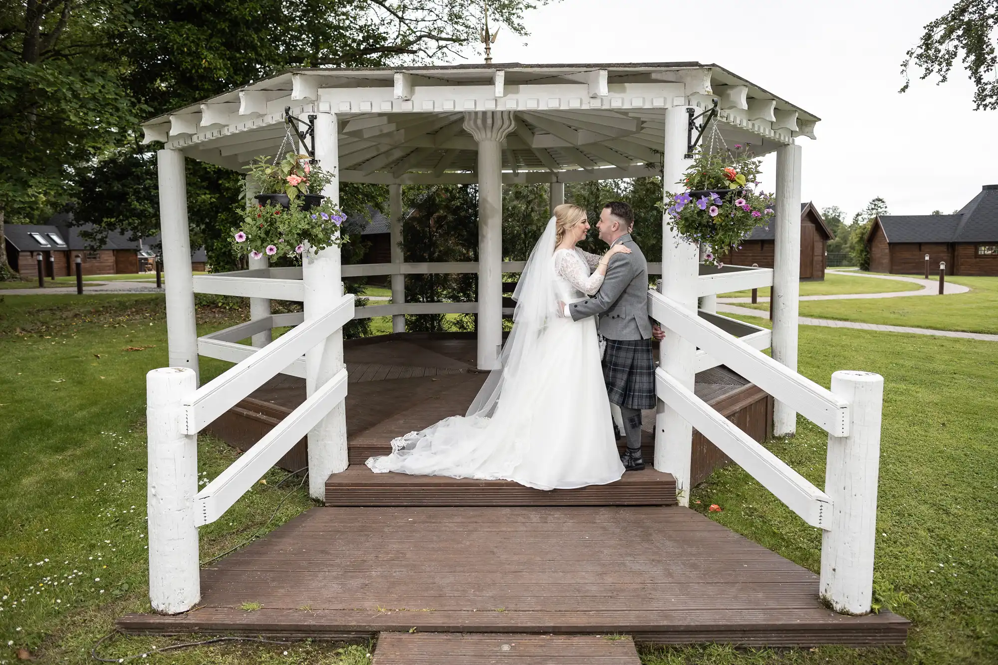 A couple in wedding attire stands under a white gazebo, sharing an intimate moment. The bride wears a long white dress while the groom is dressed in a suit with a kilt. Green grass and buildings are in the background.