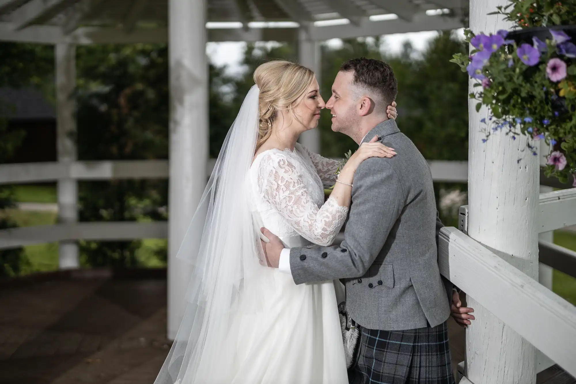 A bride and groom in a gazebo share an intimate moment. The bride wears a white lace dress and veil, while the groom is dressed in a grey jacket and patterned kilt. .