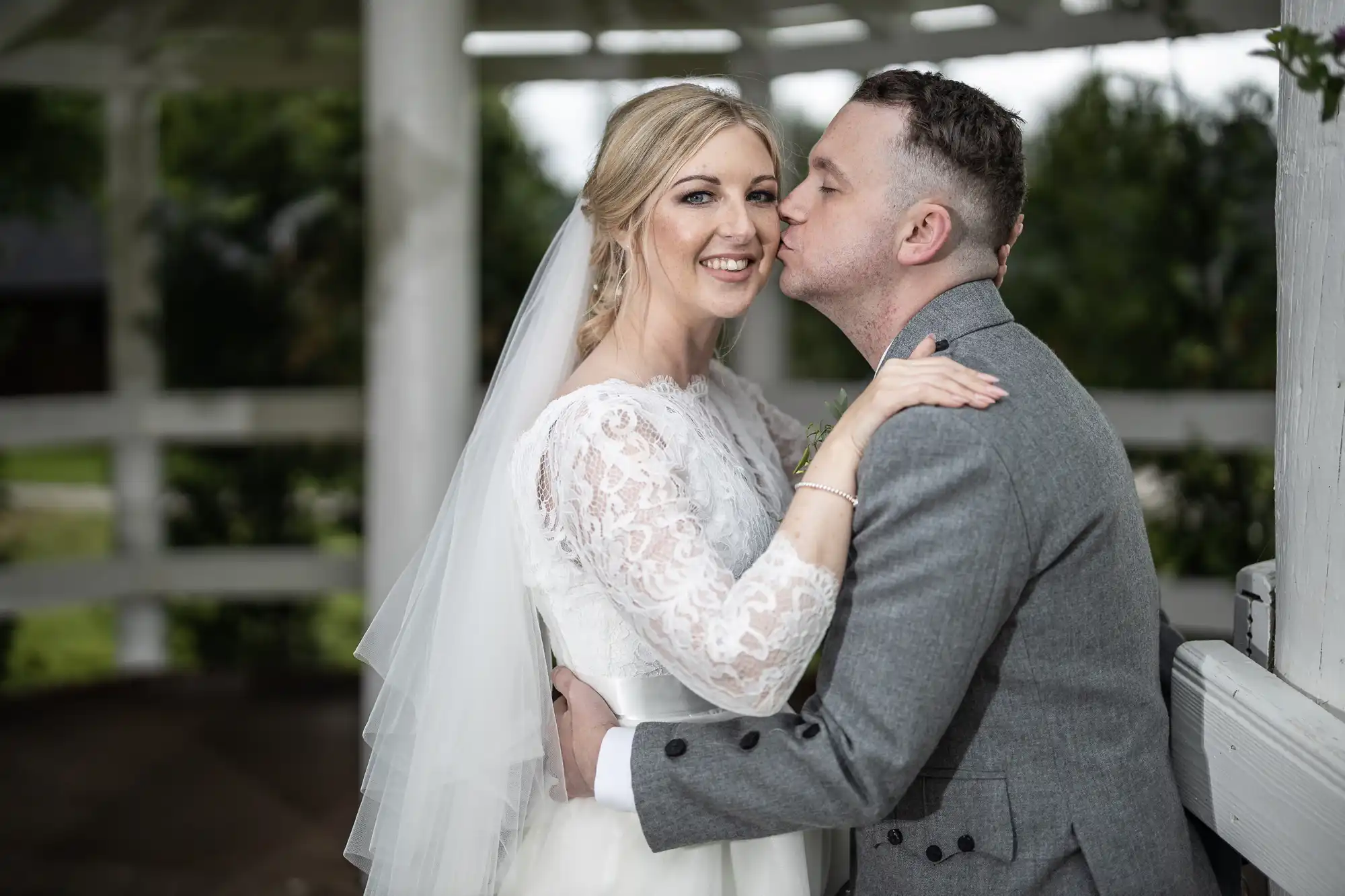 A bride in a white dress and veil smiles at the camera while the groom in a gray suit kisses her on the cheek. They are standing outdoors under a white gazebo.