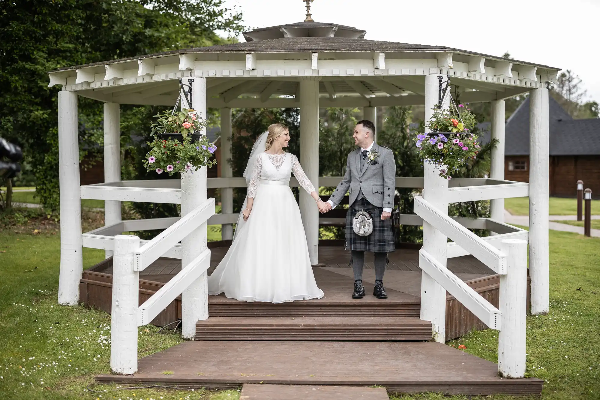 A couple stands hand in hand under a white gazebo. The woman is in a white wedding dress and the man is in a grey jacket with a black kilt. They appear to be on a wooden platform amid a grassy area.