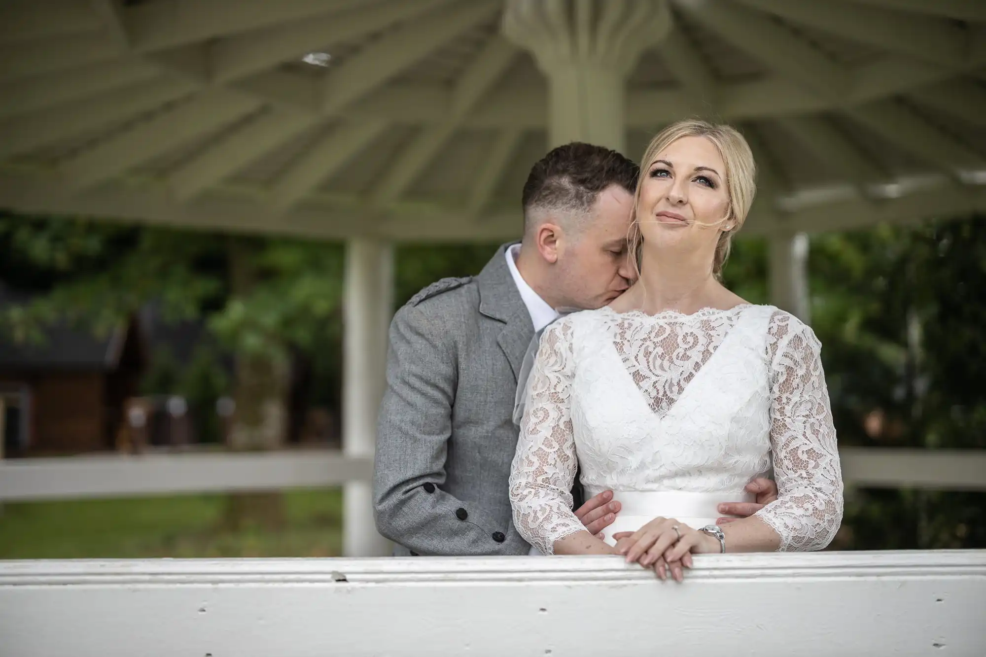 A couple standing in a gazebo, with the man in gray suit embracing and kissing the woman in a white lace dress on the neck. Both appear to be content and in a serene outdoor setting.