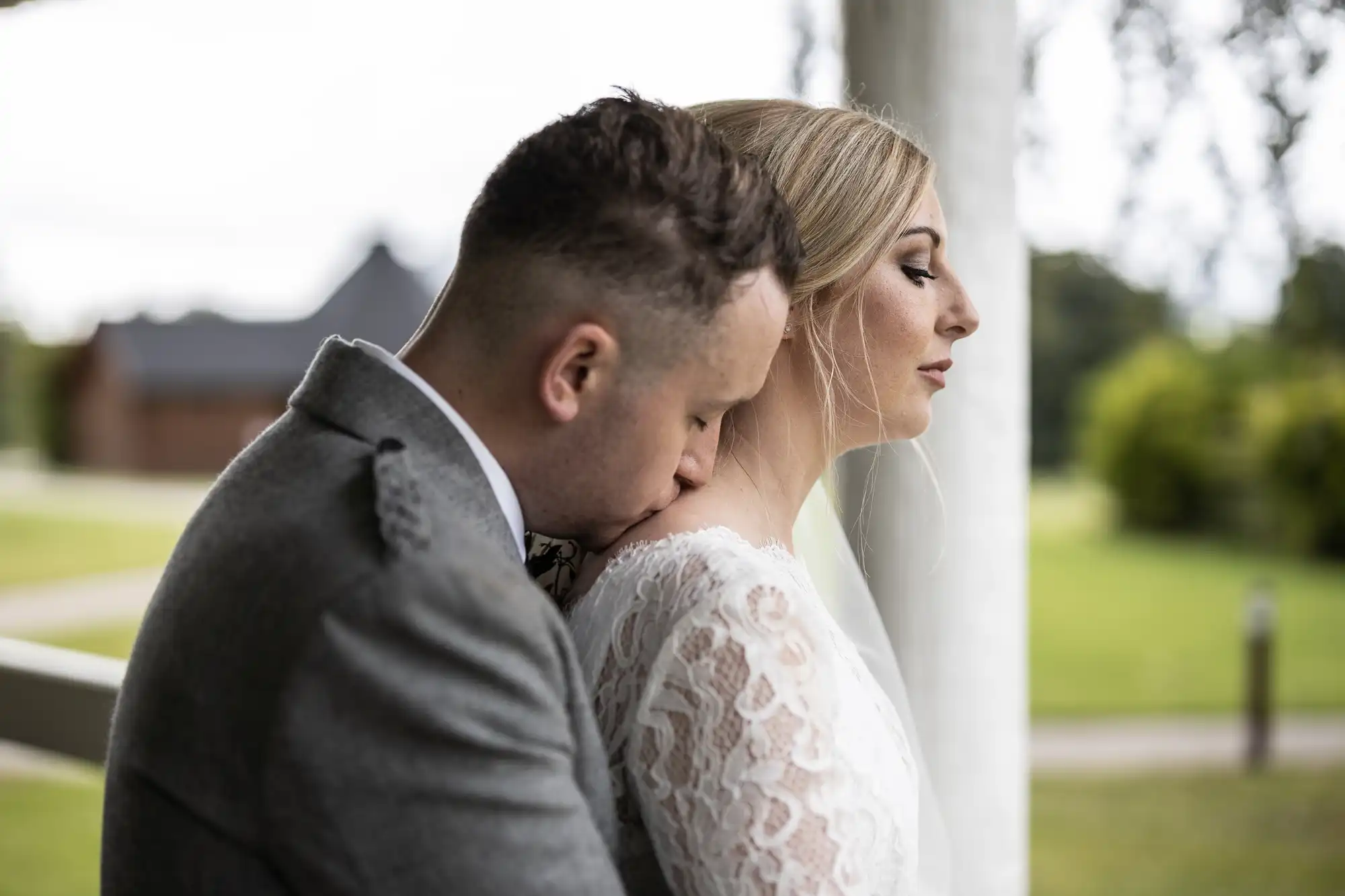 A groom in a gray suit embraces his bride, who is wearing a white lace dress, from behind while kissing her shoulder. They stand outdoors near a white pillar with greenery in the background.