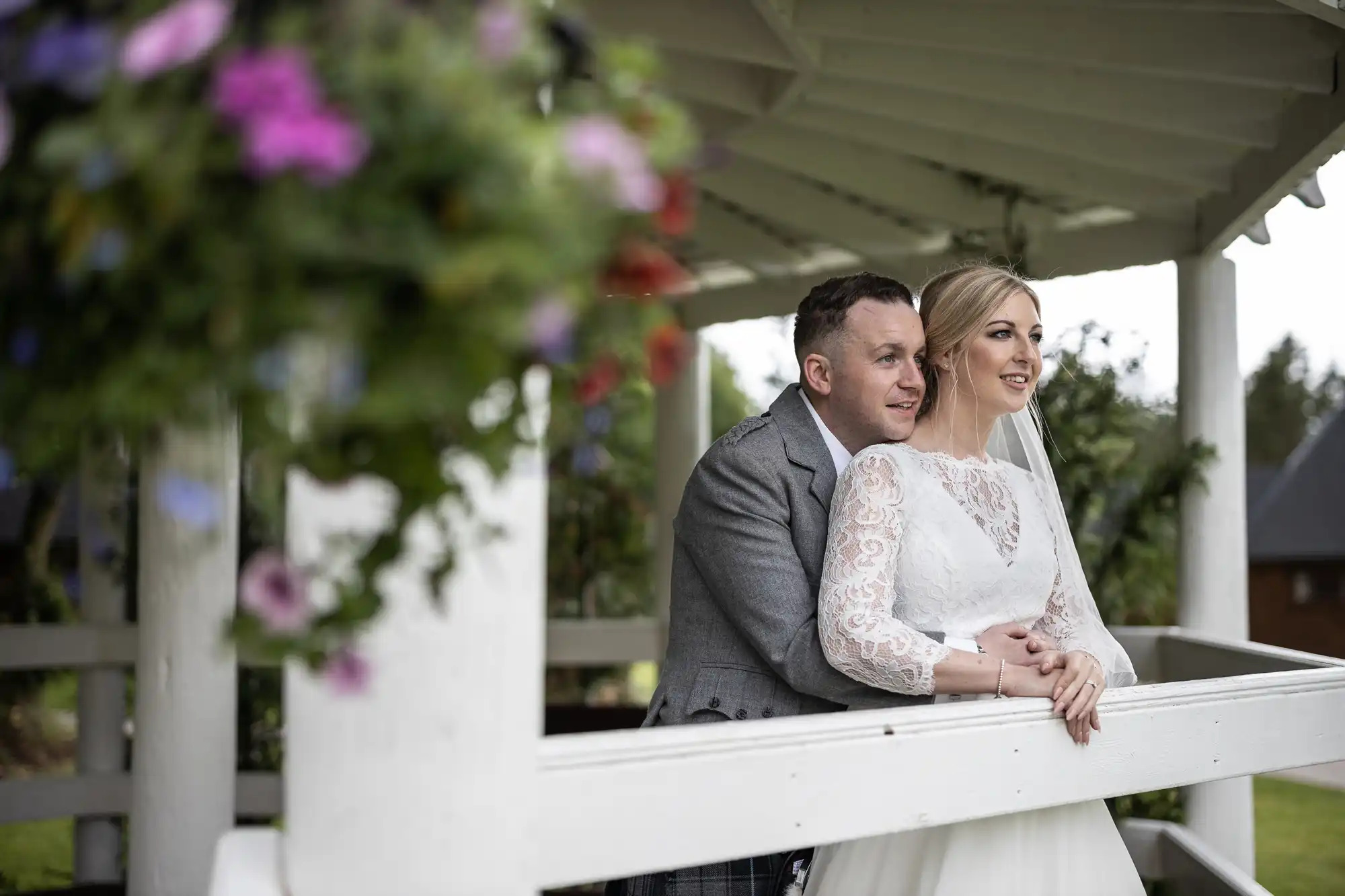 A couple in wedding attire leans on a white wooden railing under a gazebo. The bride wears a lace dress and veil, while the groom is in a gray suit. Flowers hang in the foreground.