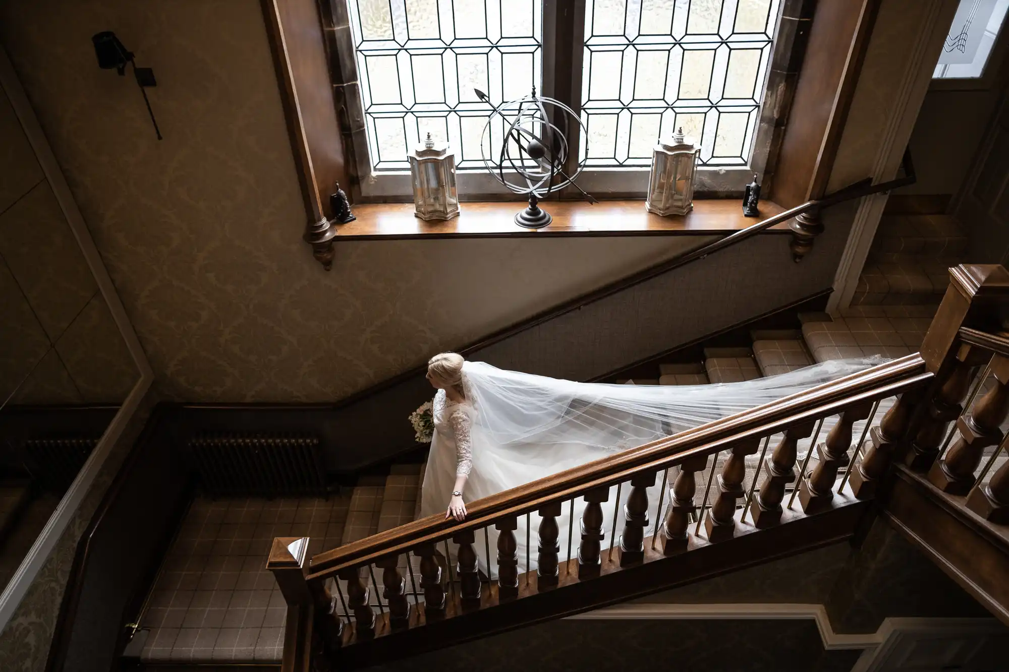 A bride in a white wedding dress and long veil descends a wooden staircase with large windows, holding a bouquet.