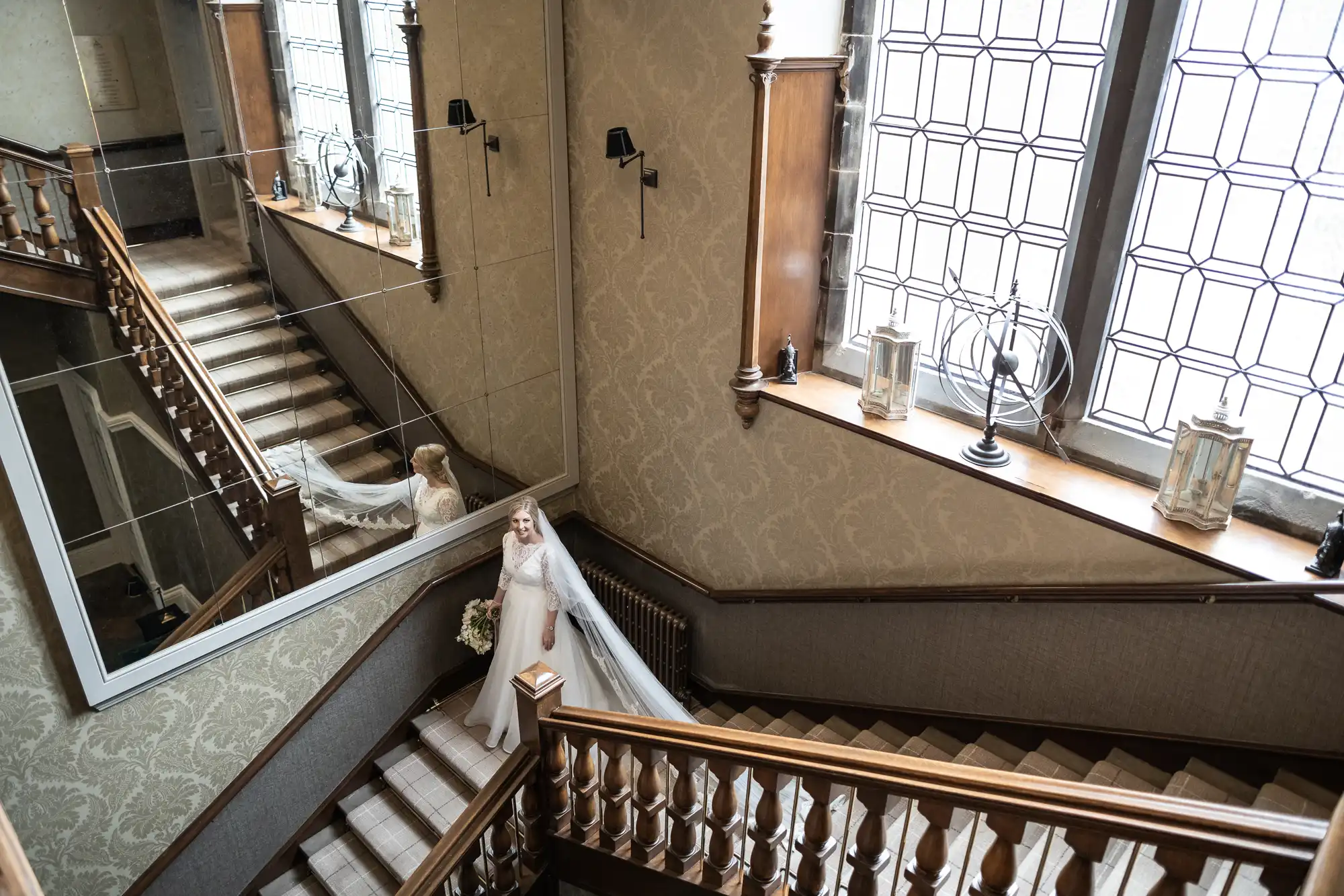 Bride in a white gown descends a grand wooden staircase with large windows and mirrors reflecting her image.