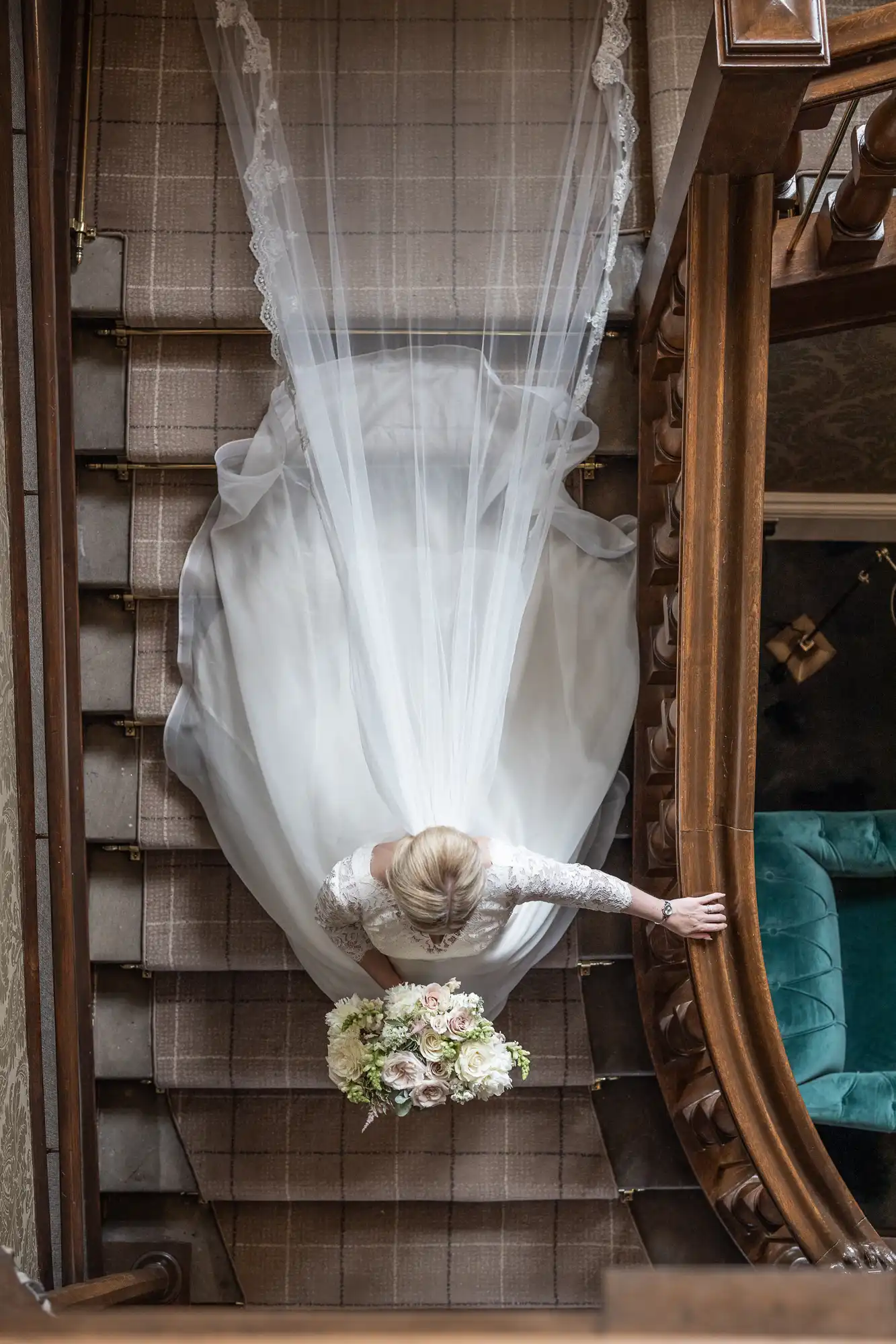 A bride in a flowing white dress and veil is seen from above, holding a bouquet of flowers while descending a staircase.