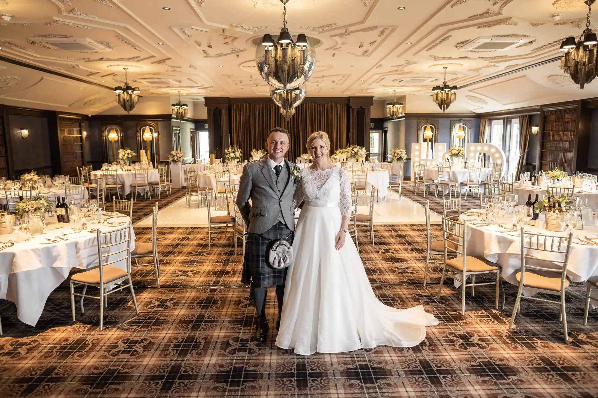 A couple dressed in wedding attire stands in the center of an elegantly decorated banquet hall with set tables and large illuminated initials in the background.