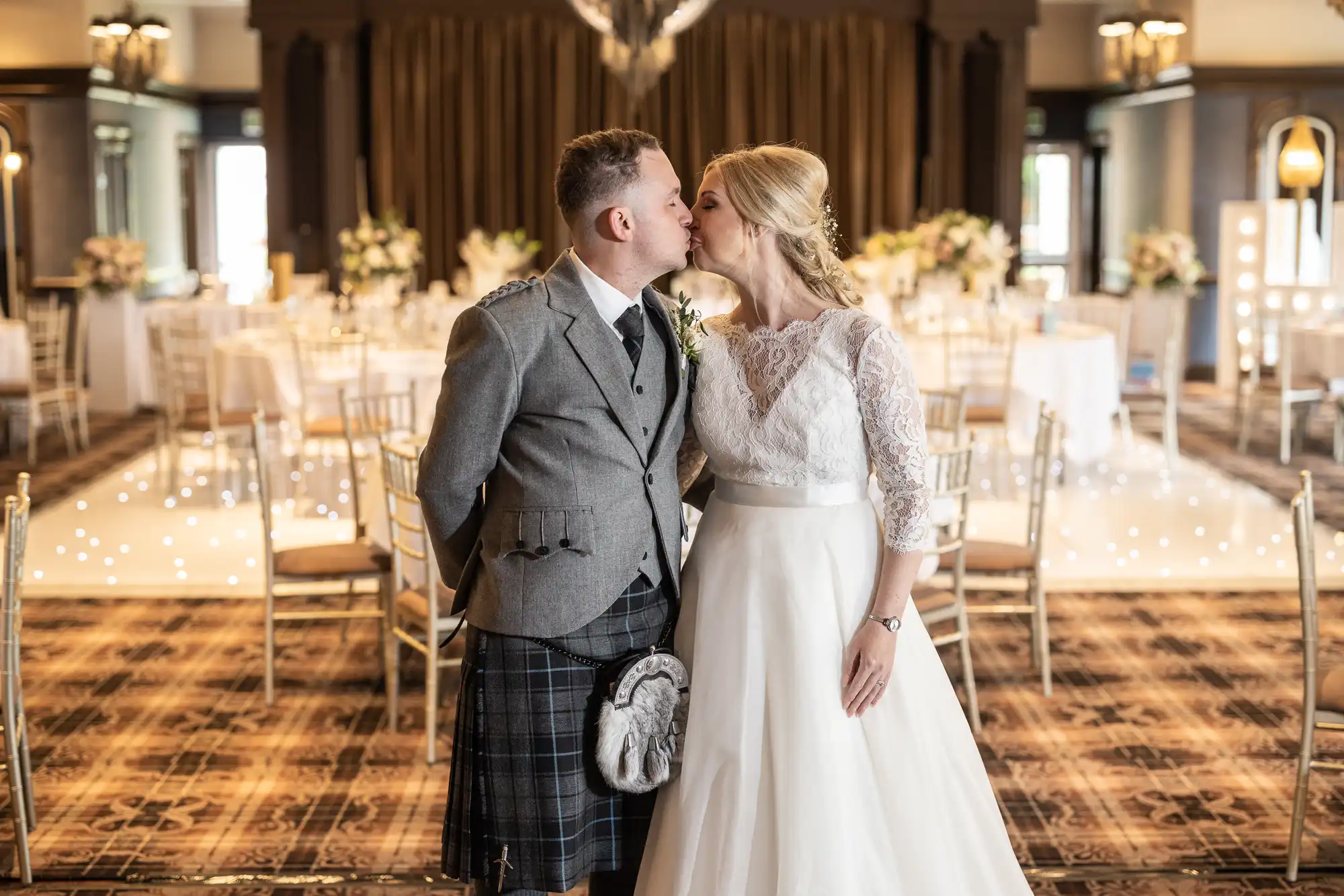 A bride and groom kiss in a well-decorated reception hall. The groom wears a grey jacket and kilt, and the bride wears a white wedding dress. Tables and chairs are arranged in the background.