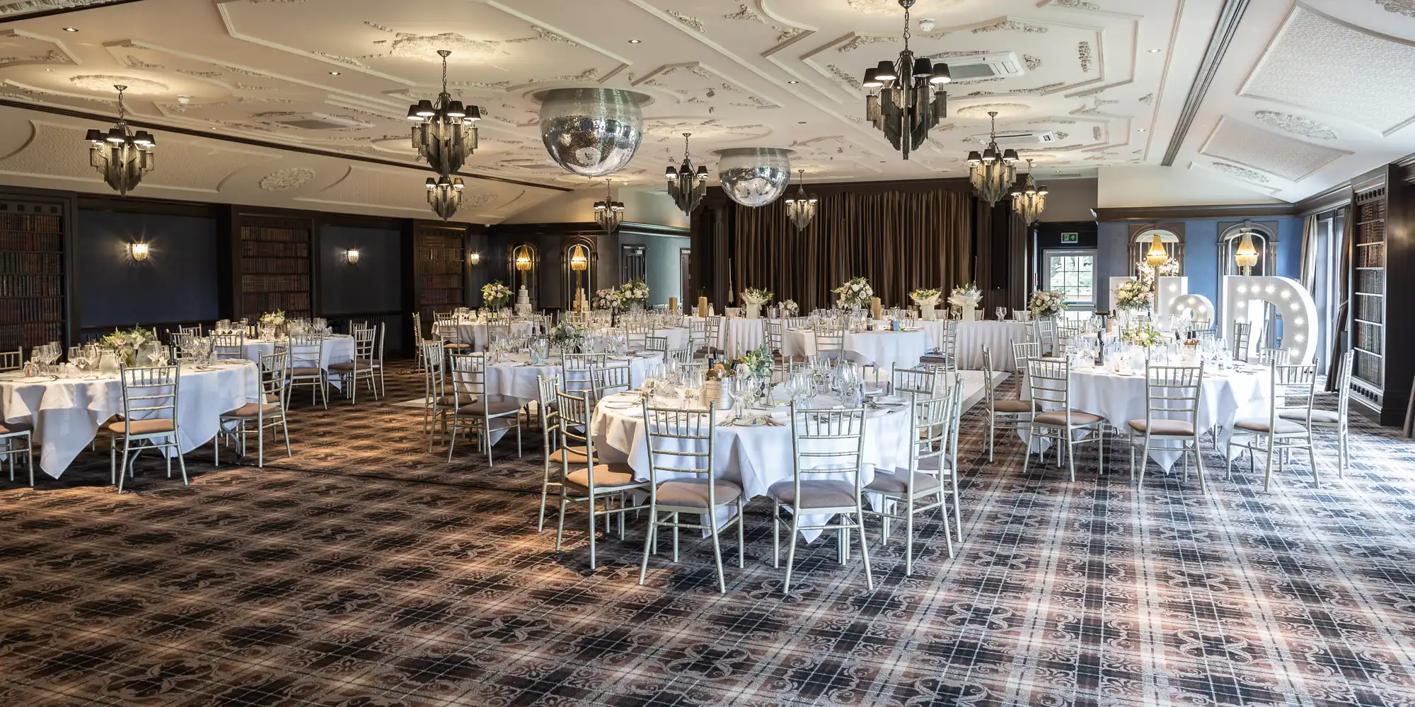 A decorated banquet hall with round tables and white chairs arranged for an event. Table settings include white tablecloths, glassware, and floral centerpieces. Chandeliers and large mirrors hang from the ceiling.