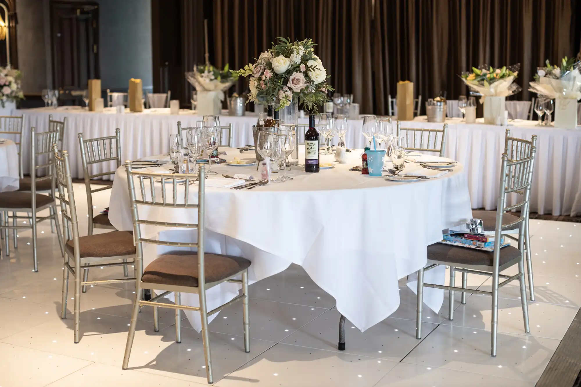 A dining table set for an event with white tablecloth, floral centerpiece, wine bottle, and tableware, surrounded by silver chairs. Additional decorated tables are in the background.