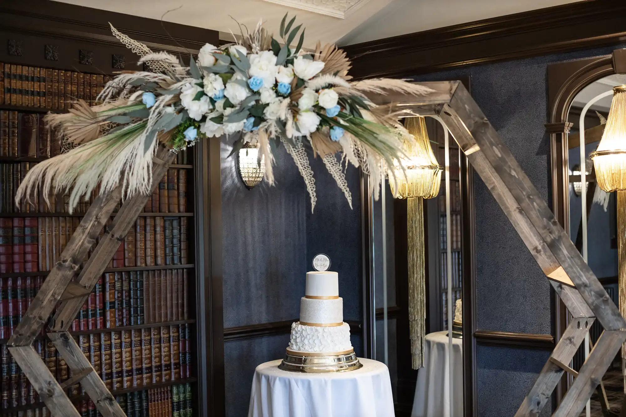 A three-tiered white cake with gold accents on a round table, under a wooden arch adorned with white and blue flowers and greenery, set in a room with dark-paneled walls and bookshelves.