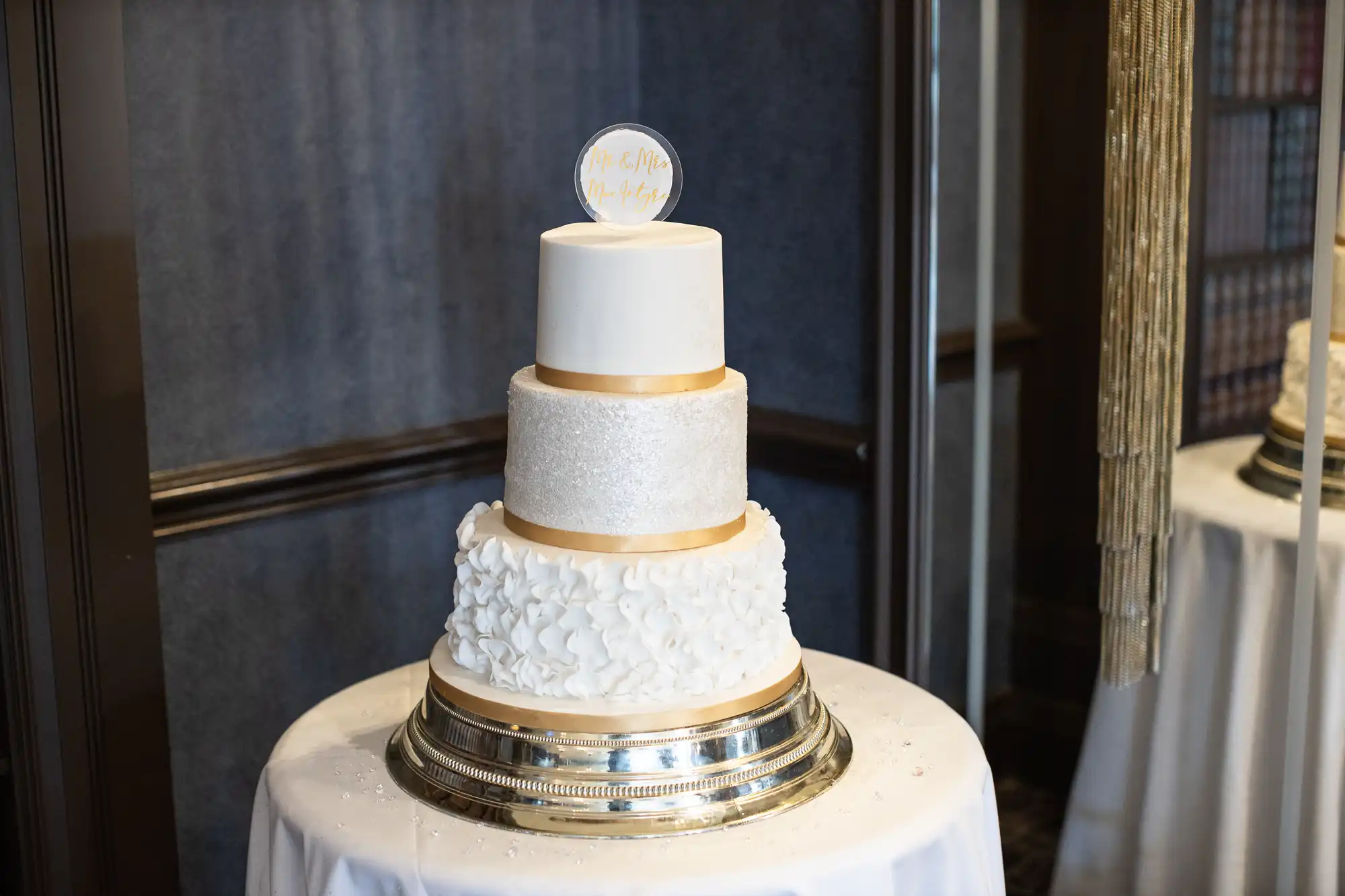 A three-tiered wedding cake with white frosting and gold accents, topped with a round sign that reads "Mr. & Mrs." This cake sits on a silver stand and table covered with a white tablecloth.