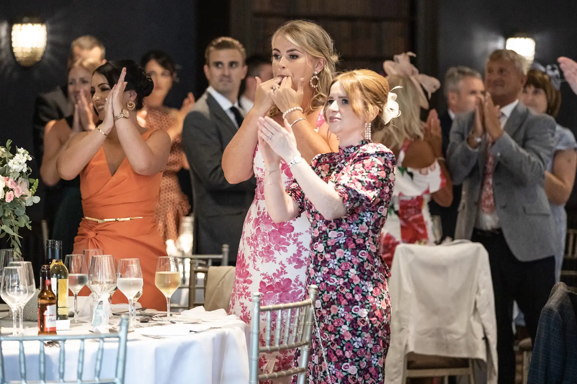 A group of people is standing and clapping in a dimly lit room, likely at an event. Two women in colorful dresses stand by a table with glassware and bottles, visible in the foreground.