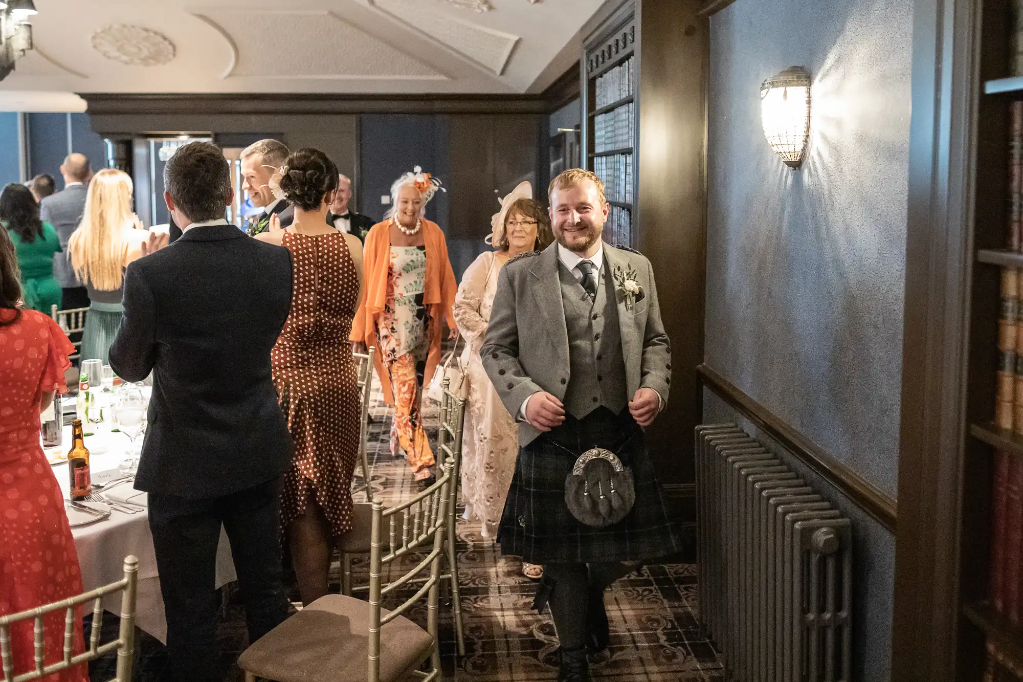 A man in a kilt leads a procession of well-dressed adults into a banquet hall with tables set for a formal event.