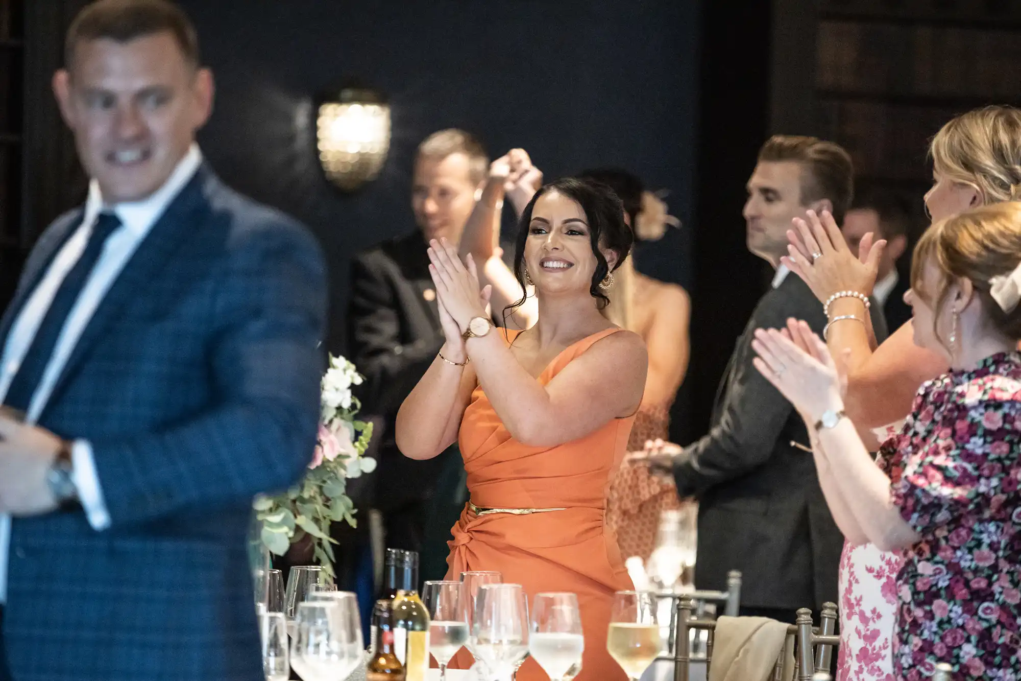 A group of people standing and clapping at an indoor event. A woman in an orange dress is in focus, appearing happy and animated.