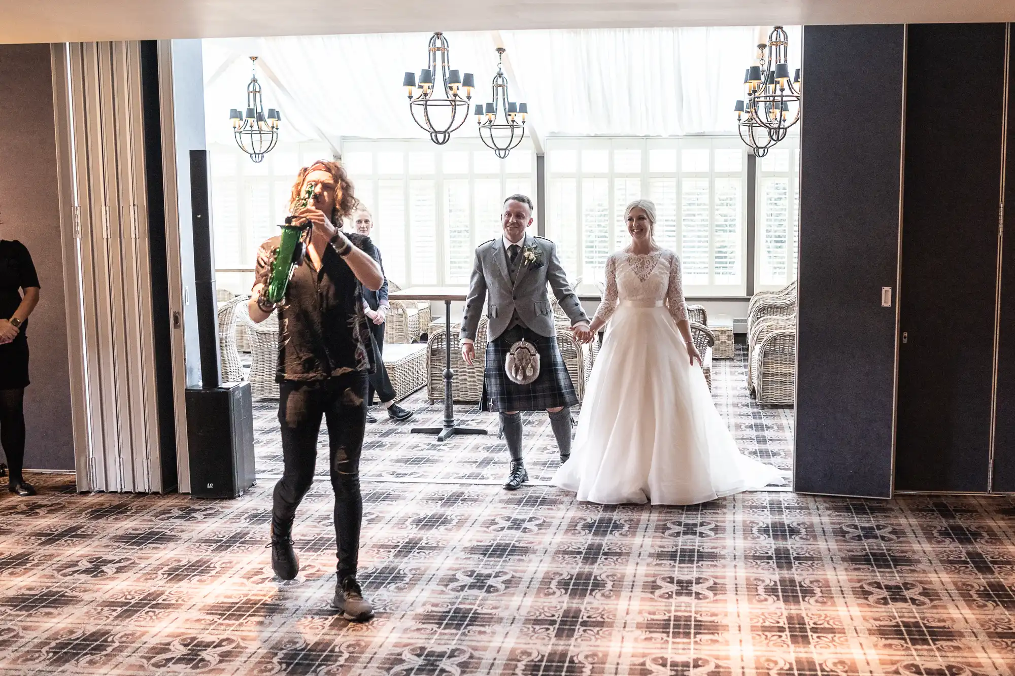 A musician leads a bride in a white gown and a groom in a kilt into a well-lit room with patterned carpet and chandeliers.