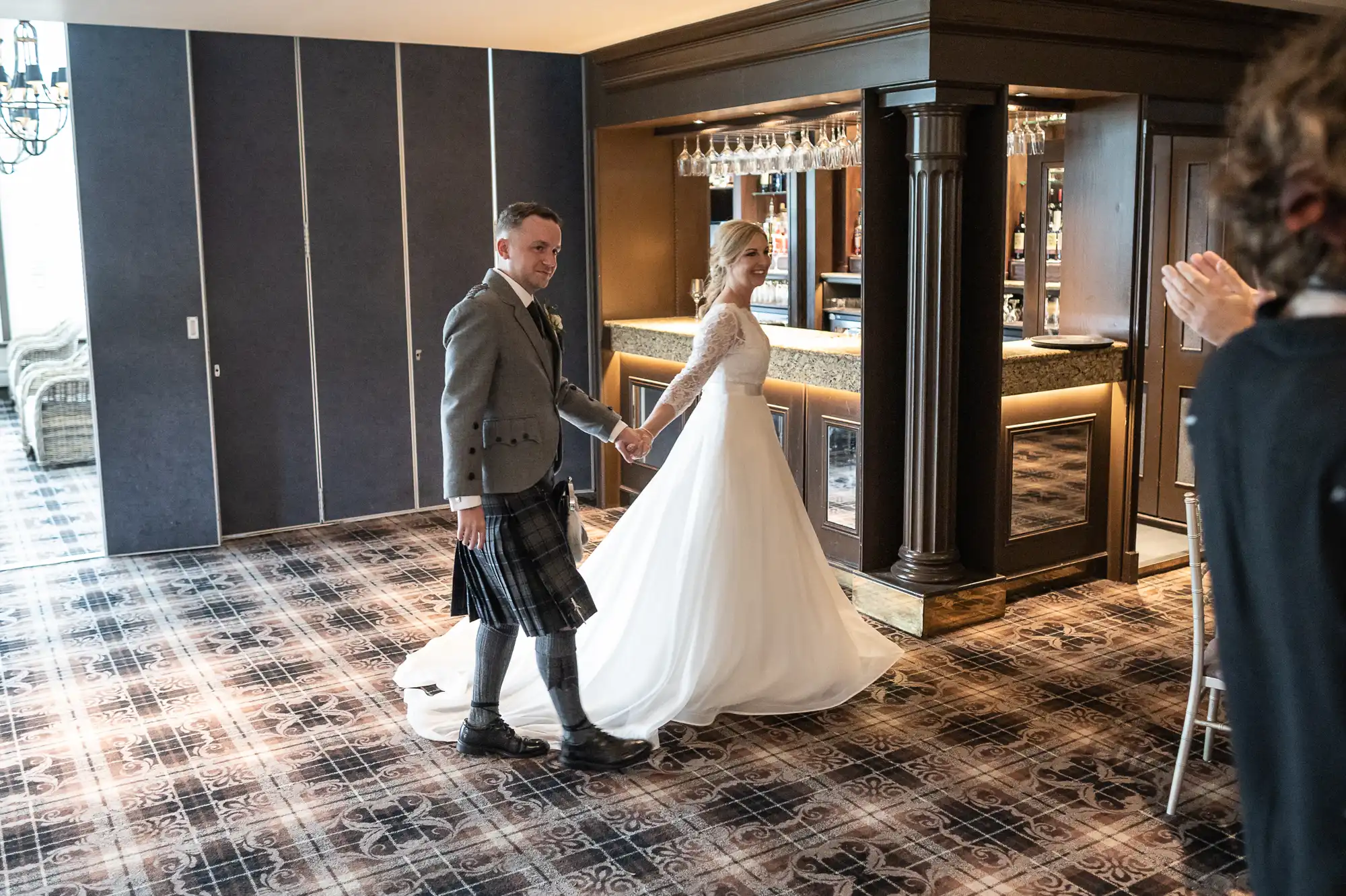 A bride and groom holding hands enter a reception hall. The groom wears a kilt, and the bride is in a white gown. Guests, slightly blurred, can be seen in the foreground applauding.