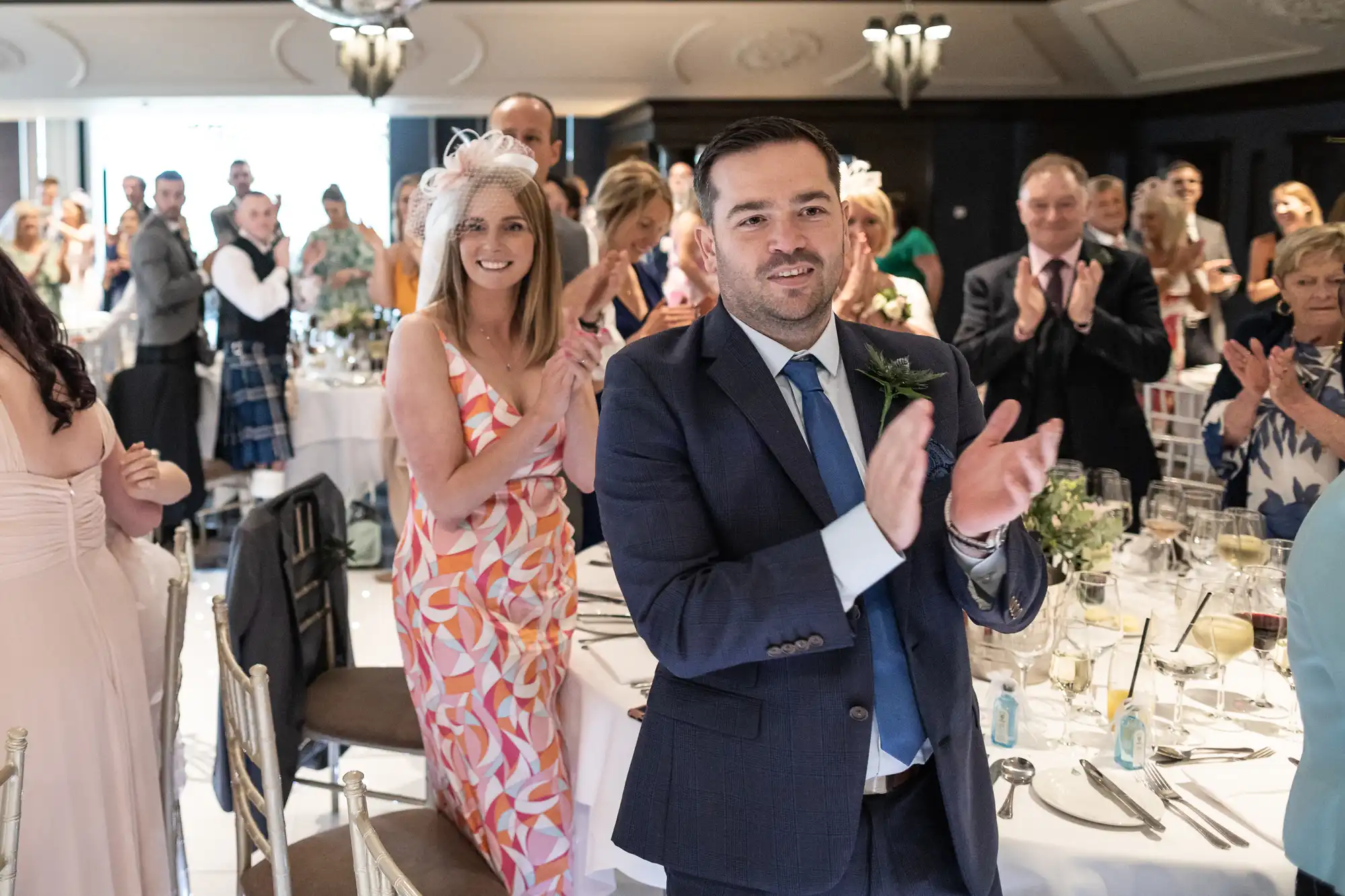 Guests at an event stand and clap. The man in the foreground wears a navy suit and the woman behind him wears a patterned dress with a white fascinator. Tables are set and decorated in the background.