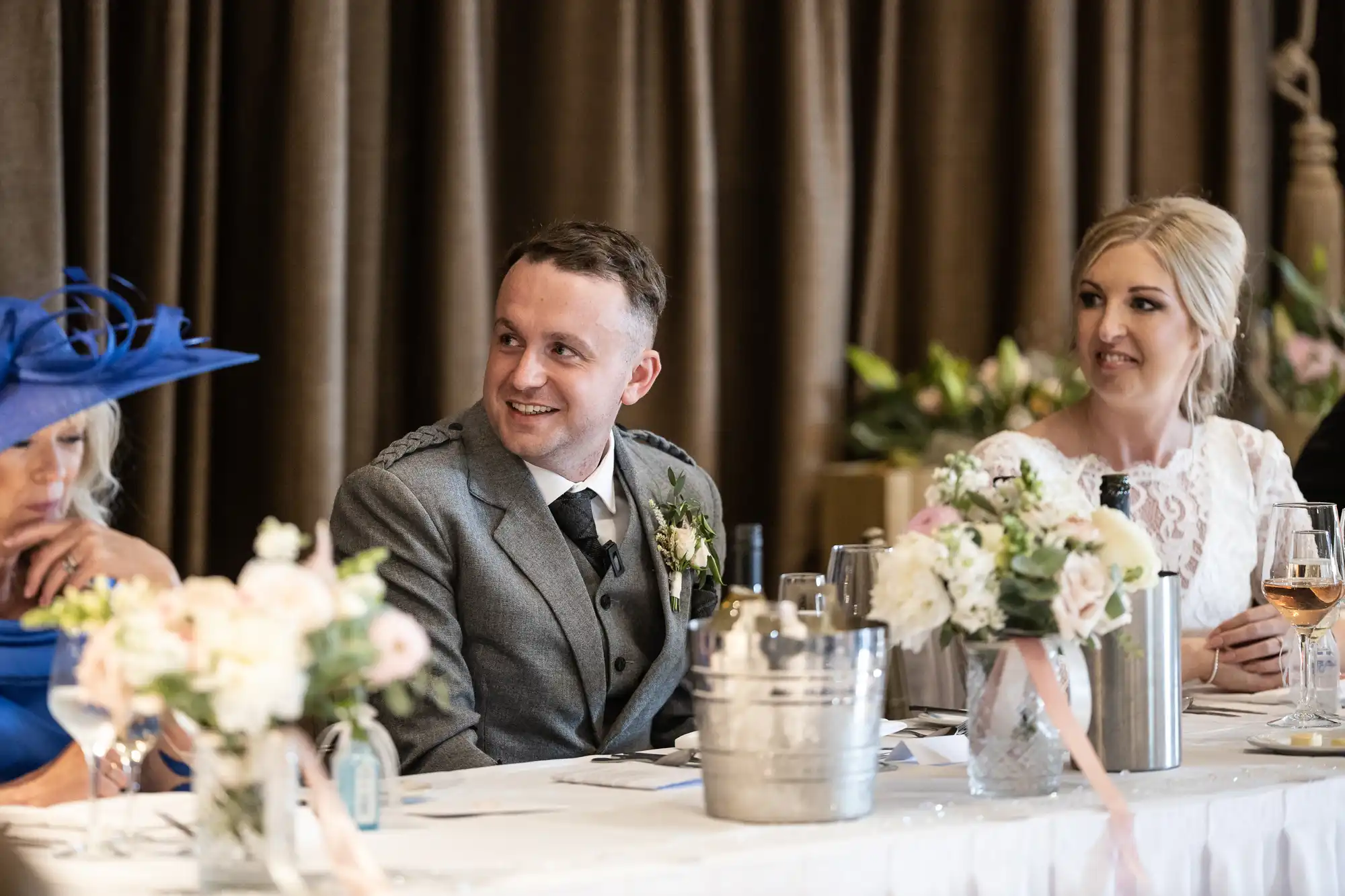 A man in a gray suit and a woman in a white dress sit at a decorated table, smiling during a formal event. Another person in a blue outfit sits nearby. Flowers and drinks are on the table.