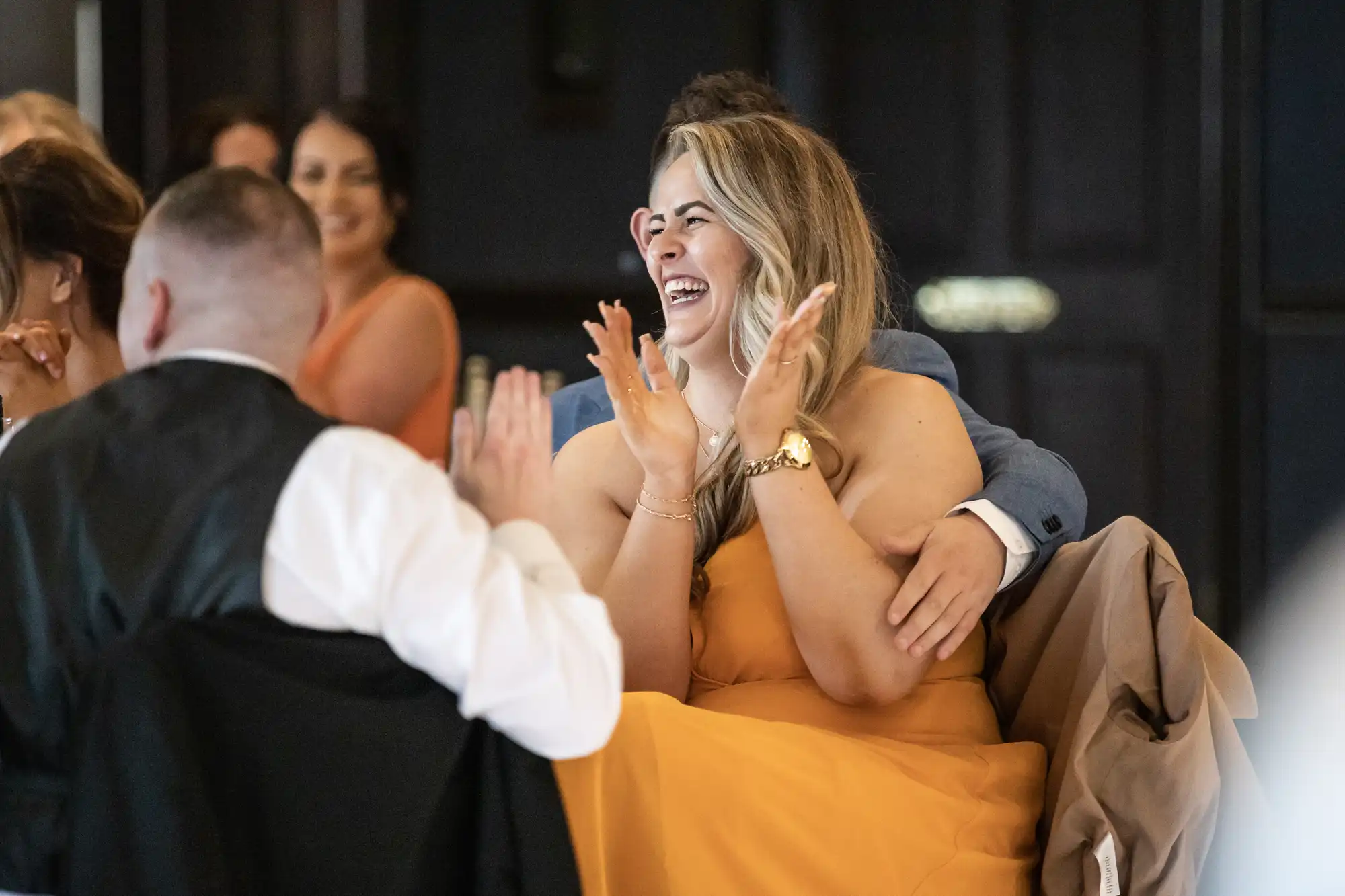 A woman in a yellow dress is smiling and clapping at an indoor event, while others nearby are seen conversing and reacting.