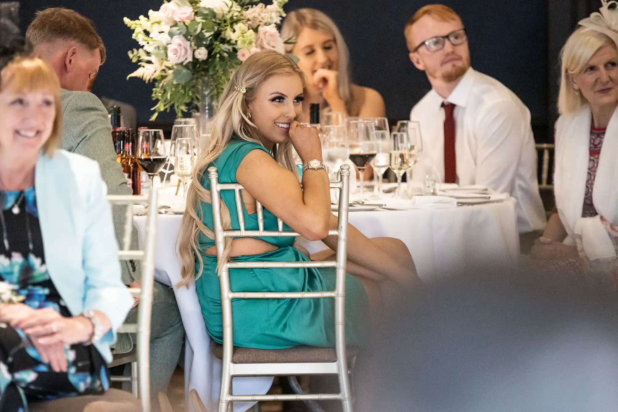 People seated at round tables at an indoor event, with a woman in a green dress in the foreground looking to her right. Tables are decorated with flowers and set with glassware.