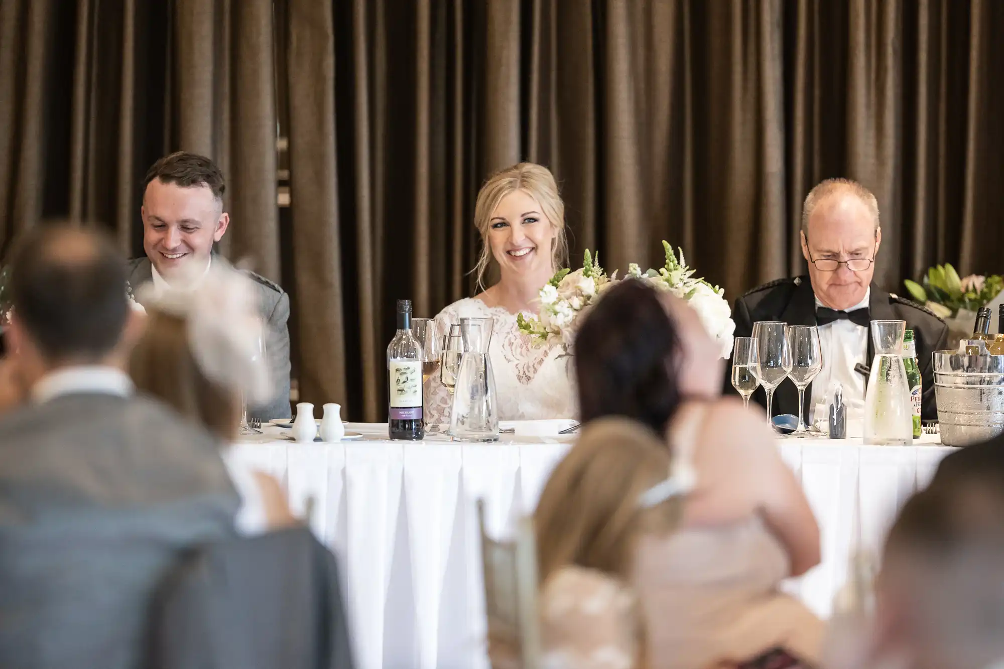 A bride and two men sitting at a draped table during a wedding reception. The bride is holding a bouquet and smiling, while guests are seated facing them.