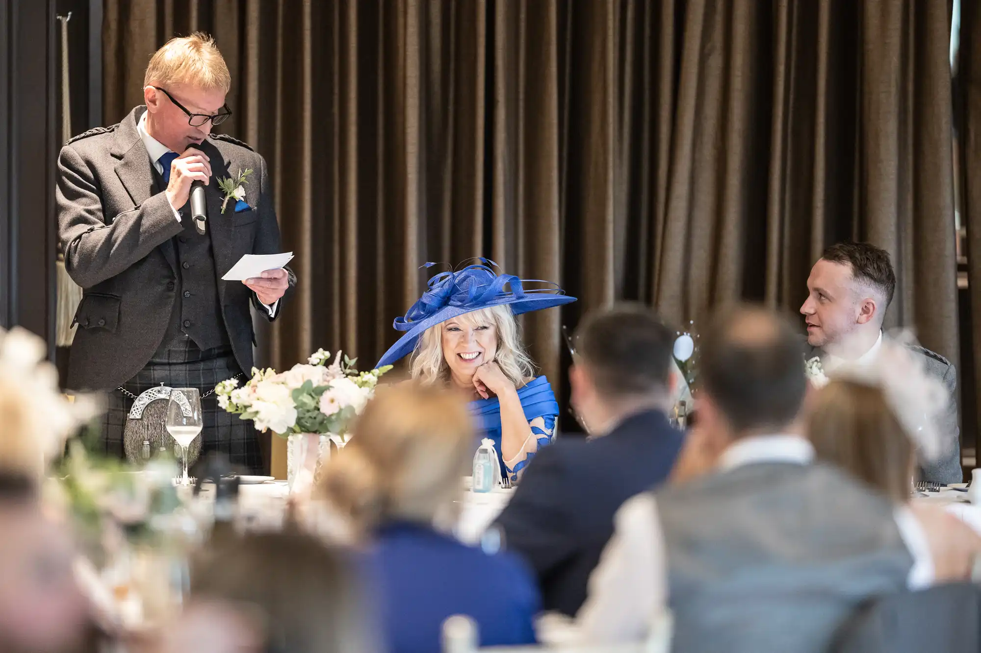 A man in a suit speaks into a microphone while holding a paper. A woman in a large blue hat and a man in a vest are seated at a table, smiling and listening. Other guests are seated around them.