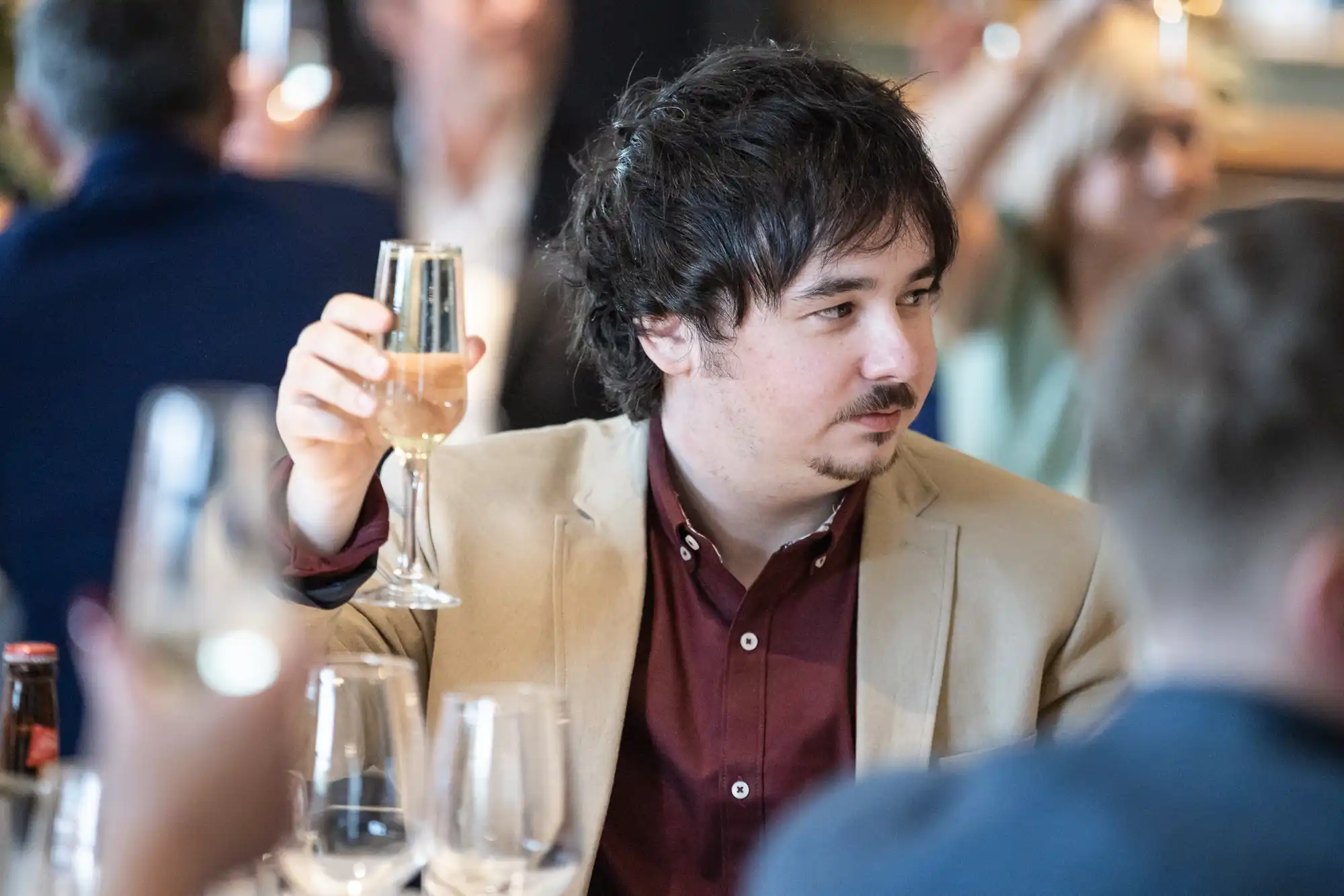 A person in a beige blazer and maroon shirt holds a champagne flute, surrounded by others in a social gathering.