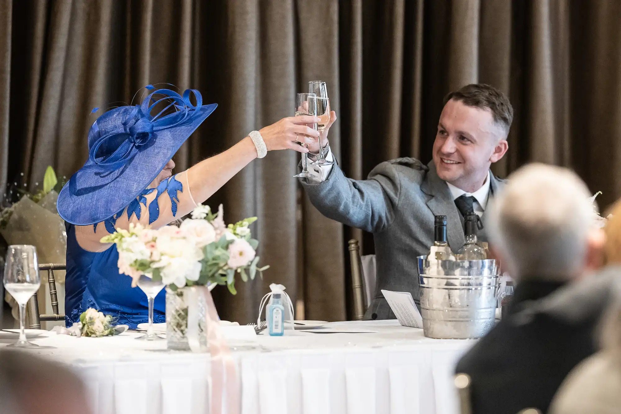 Two people dressed formally sit at a table, raising their glasses in a toast. The woman wears a blue hat and dress, while the man is in a grey suit. A small bouquet of flowers and a drink cooler are on the table.