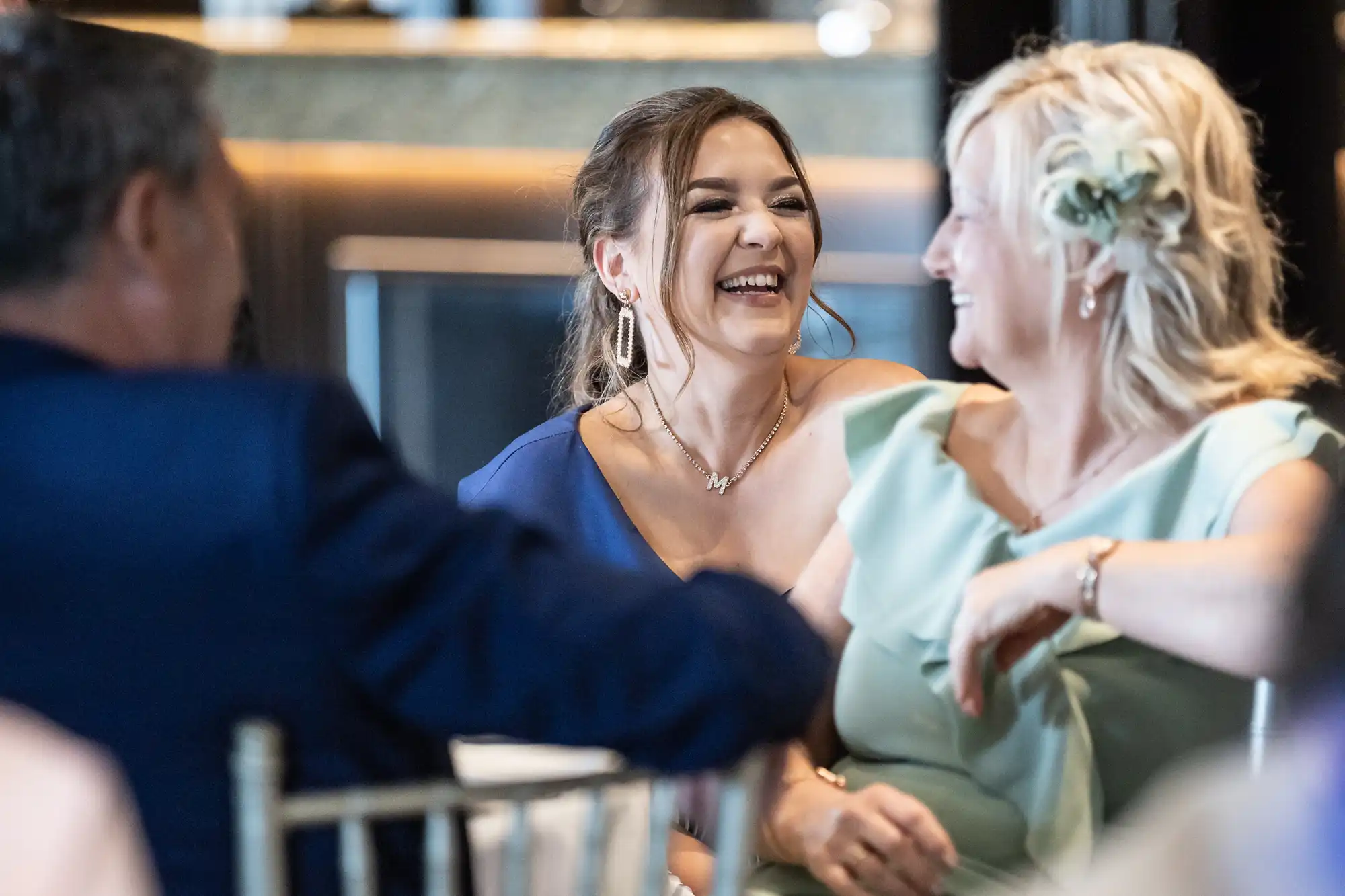 Two women smiling and laughing, one wearing a blue dress and the other a light green outfit with a flower in her hair, while a man in a suit sits next to them.