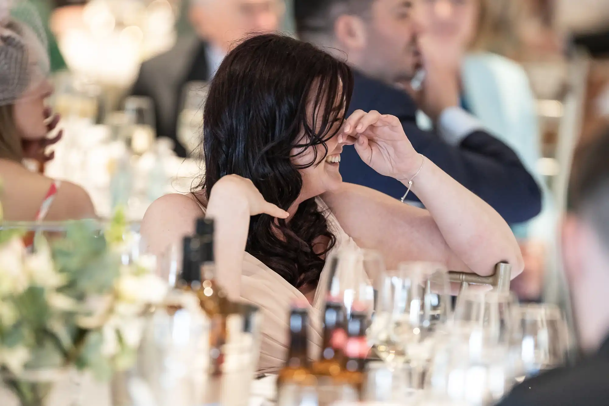 A woman with dark hair laughs, covering her face with her hand, at a formal event with others seated at tables filled with wine glasses and bottles.