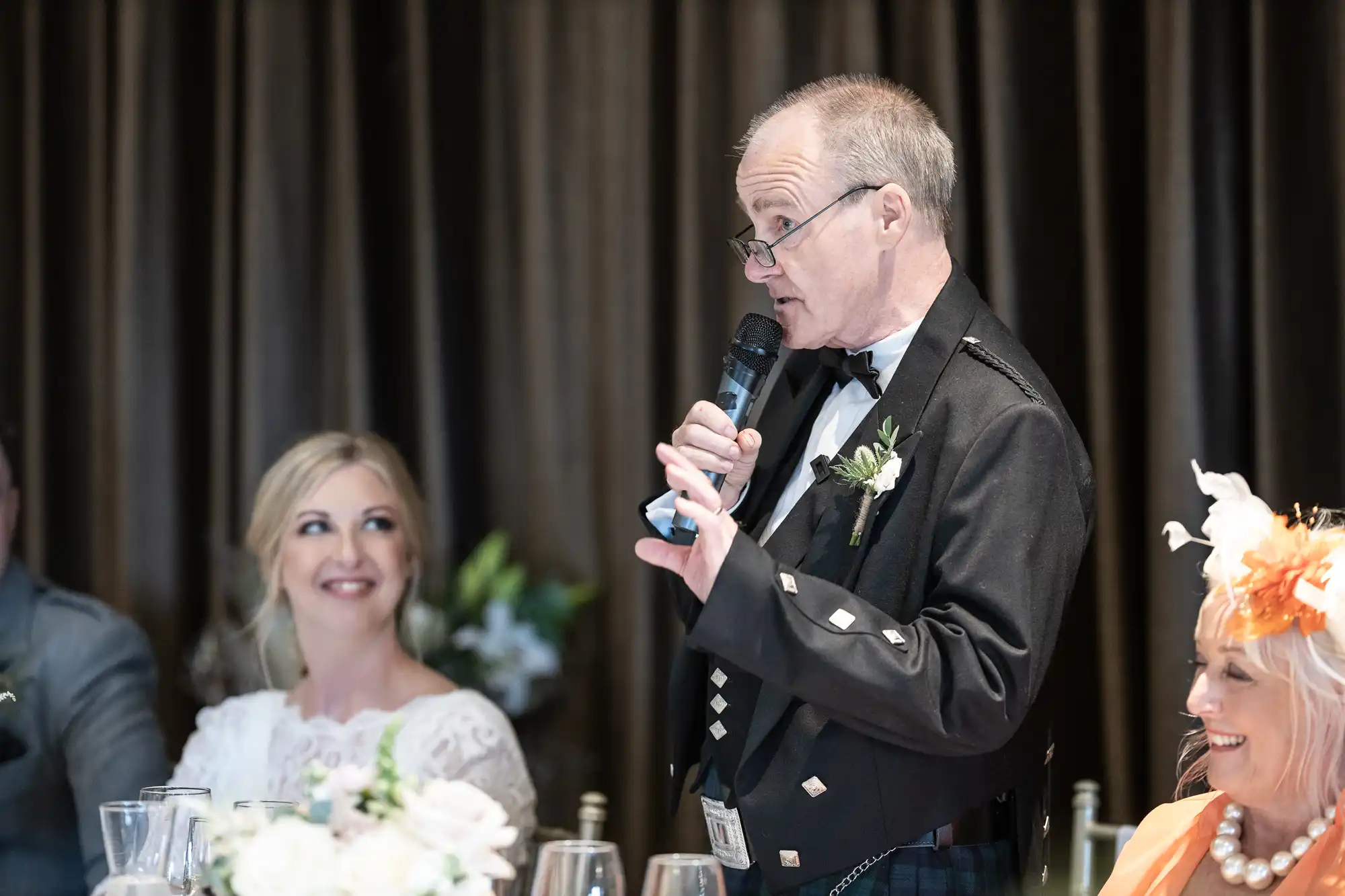 An elderly man in formal attire is giving a speech with a microphone while a woman in a wedding dress looks at him and smiles. Another person in a hat is seated nearby.