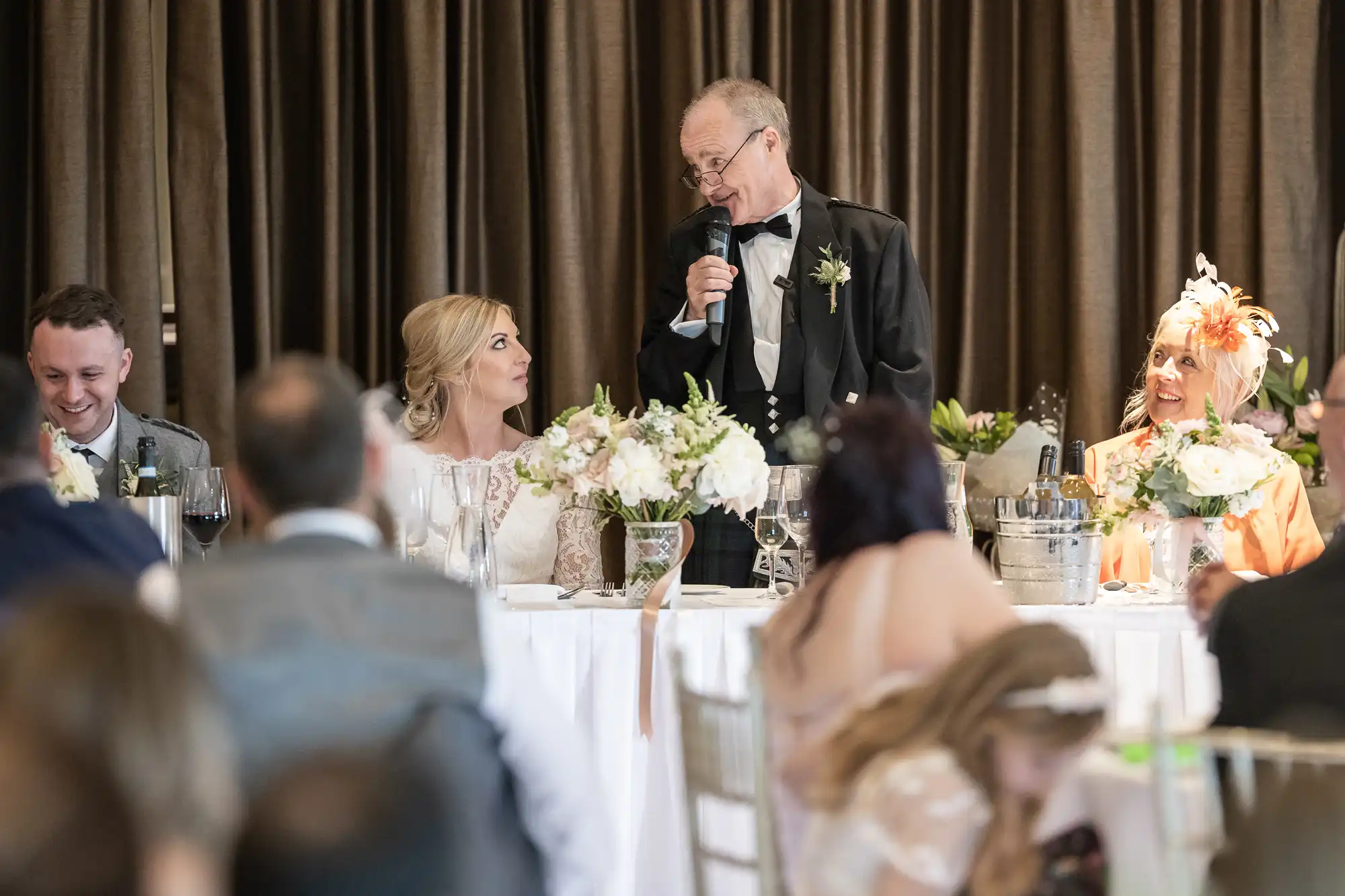 A man in a black suit is giving a speech with a microphone at a wedding reception, while seated at a table with flowers are two women and one man, all dressed in formal attire.