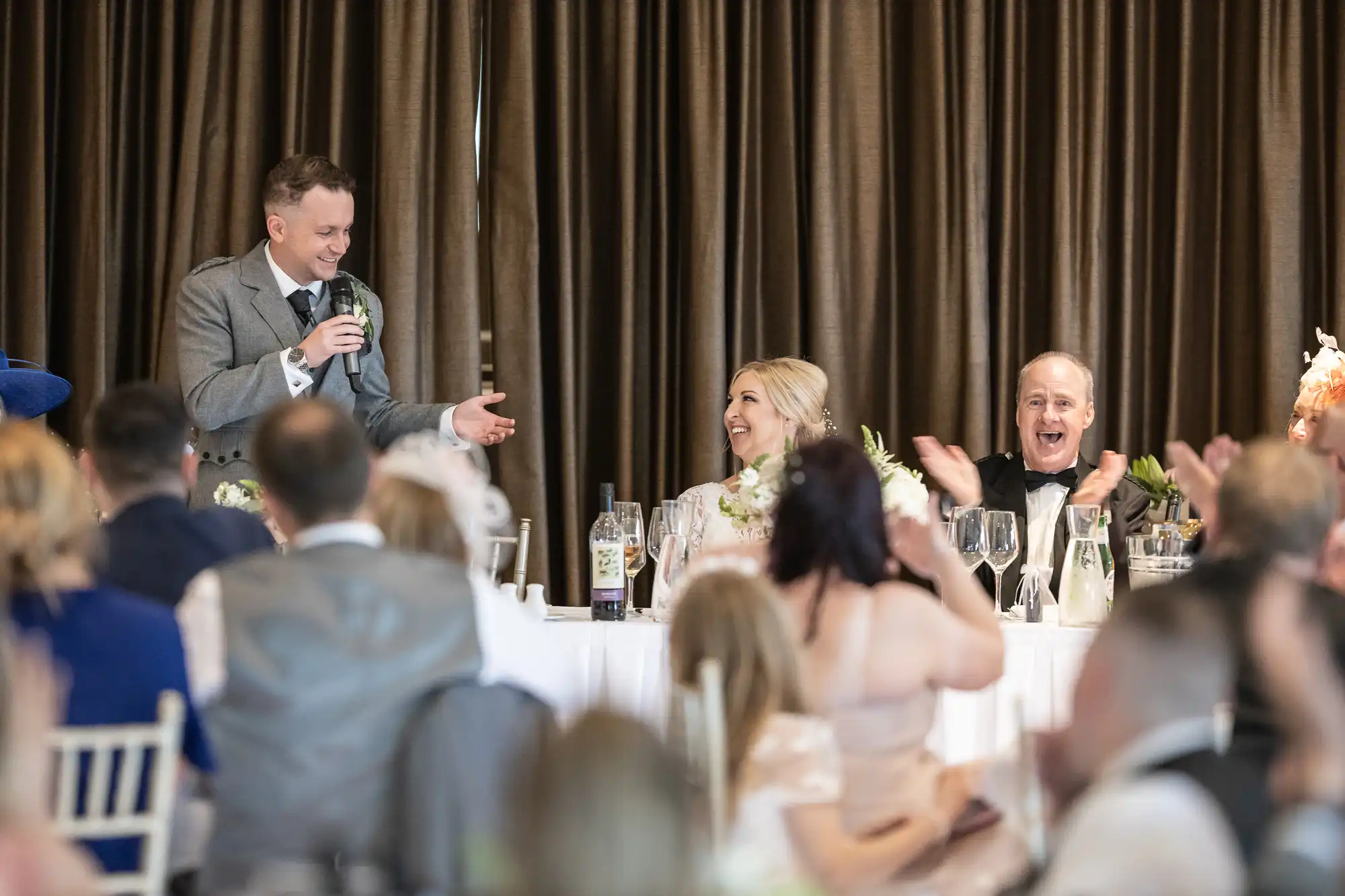 A man in a gray suit speaks into a microphone at a banquet table, while a woman in a white dress smiles and a man in a black suit applauds. Guests at the event are clapping and smiling.