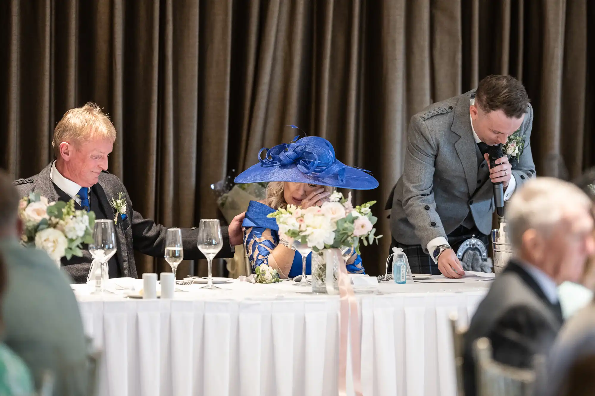 Three people at a formal event seated at a table adorned with flowers and glassware. The person on the right is speaking into a microphone, while the person in the middle covers their face with their hand.