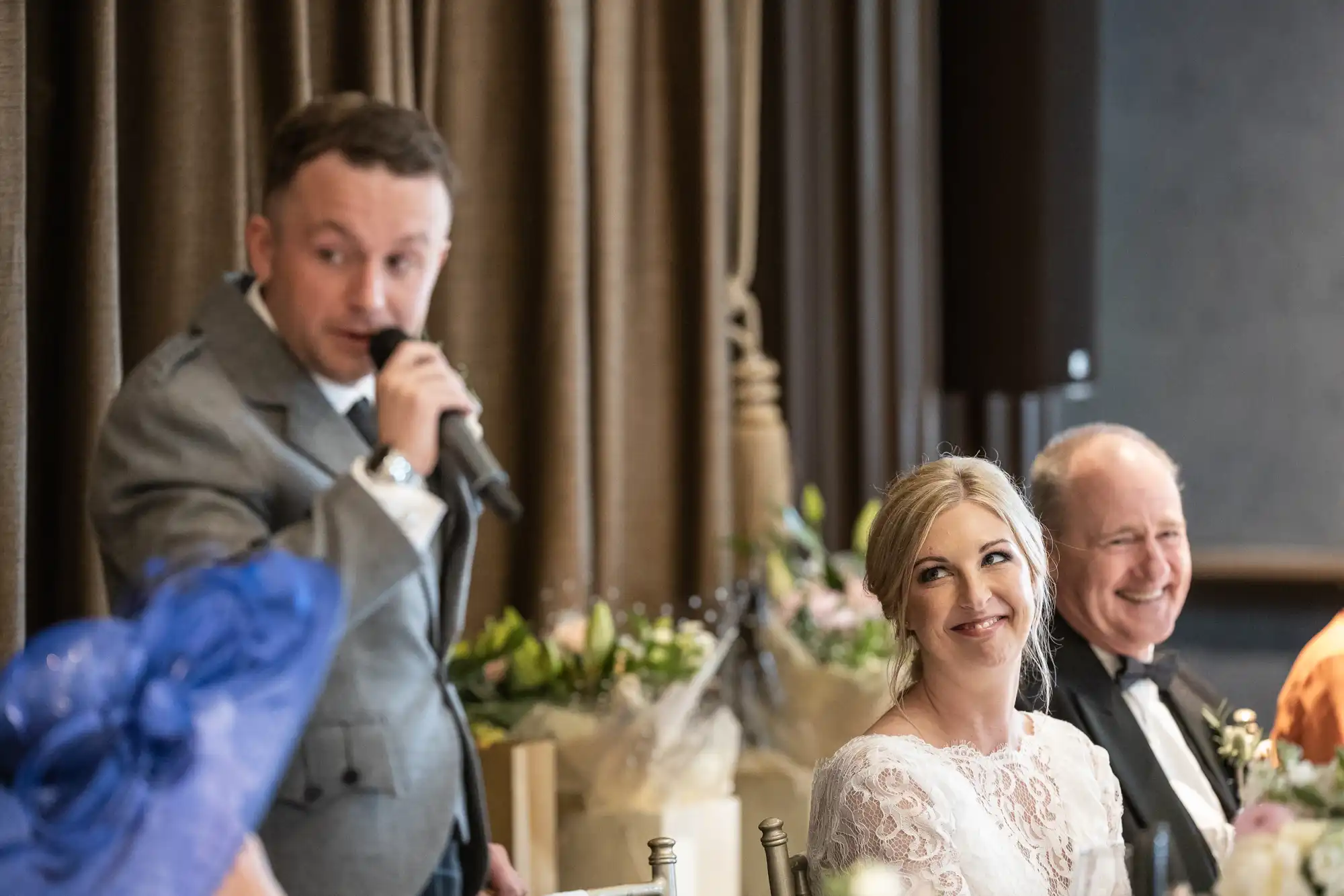 A man in a gray suit speaks into a microphone at a wedding reception while a seated bride and another man listen and smile.