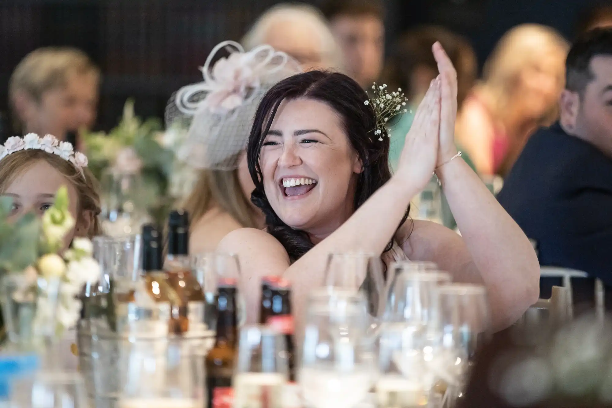 A woman with dark hair applauds and smiles while seated at a table with other guests, surrounded by bottles and glasses, during a formal gathering.