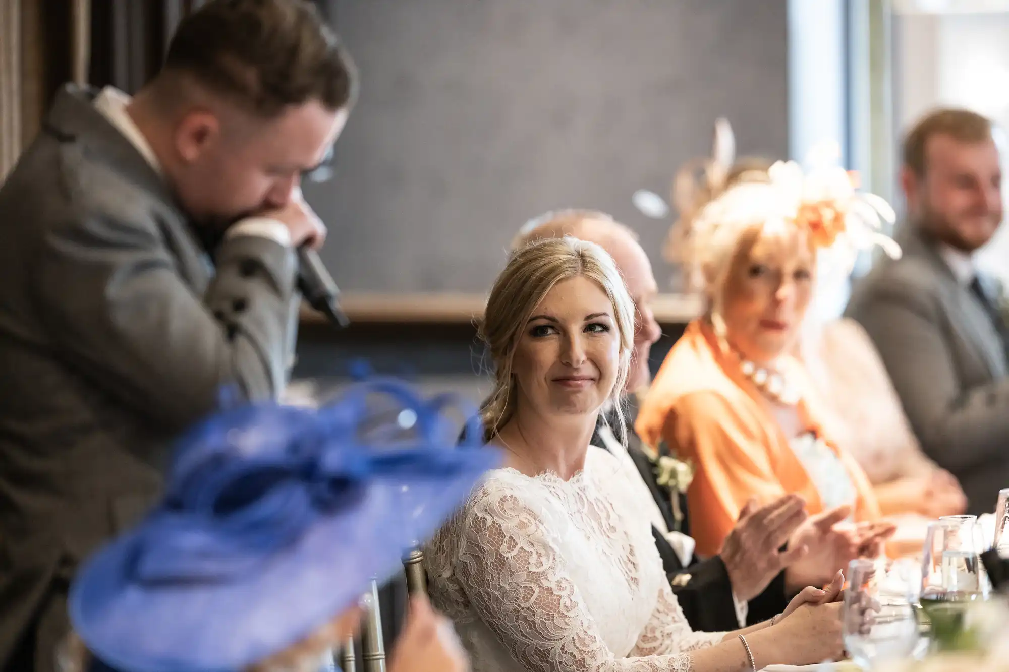 A bride in a white lace dress smiles at a man speaking into a microphone during a wedding reception. Other guests are seated, some clapping and dressed formally.