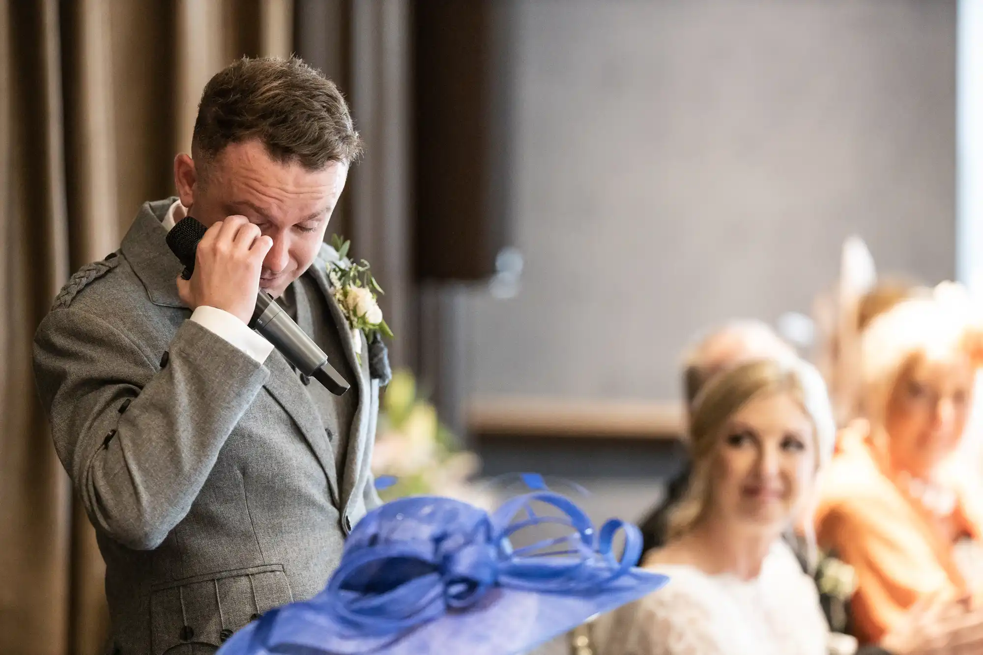 A man in a grey suit wipes a tear from his eye while speaking into a microphone. A woman in a white dress and a blue hat are seated beside him, watching attentively.