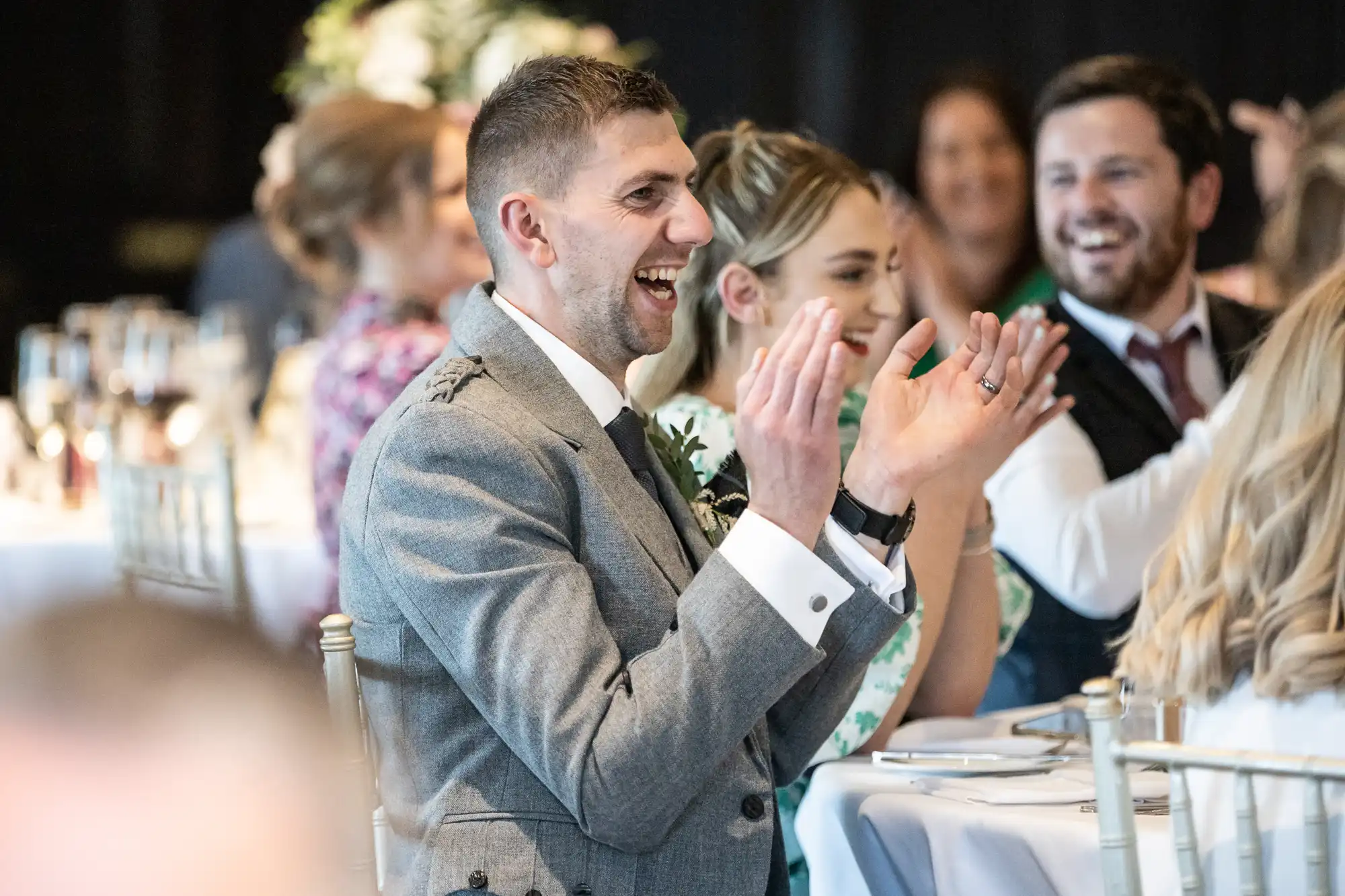 A man in a gray suit and tie smiles broadly and claps while seated at a decorated table alongside other smiling guests.