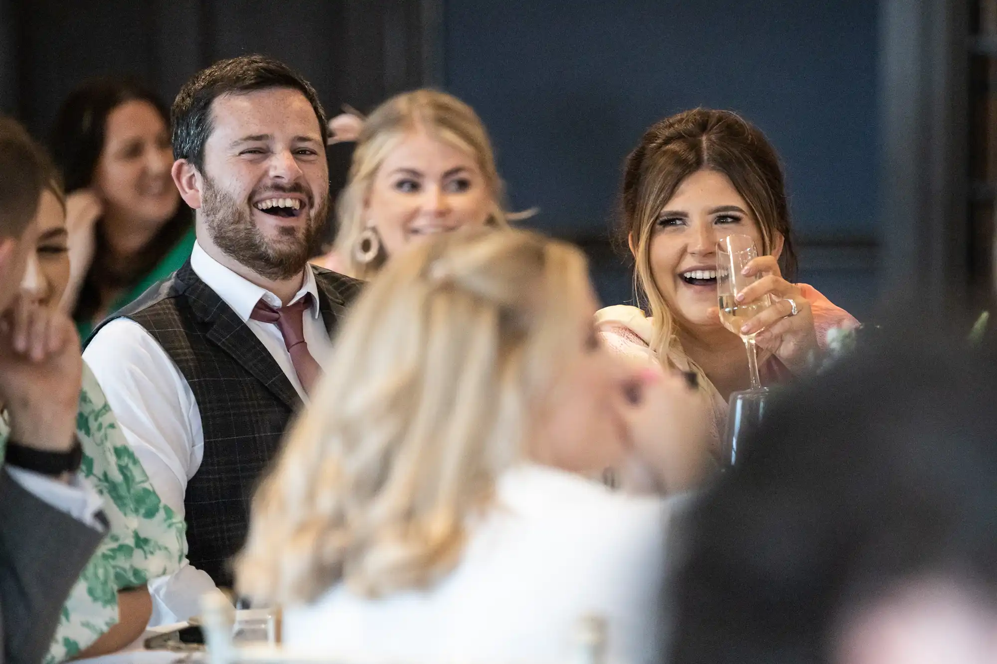 A group of people sitting at a table with a man and a woman in the foreground laughing and holding drinks. They are indoors, with a dark background.