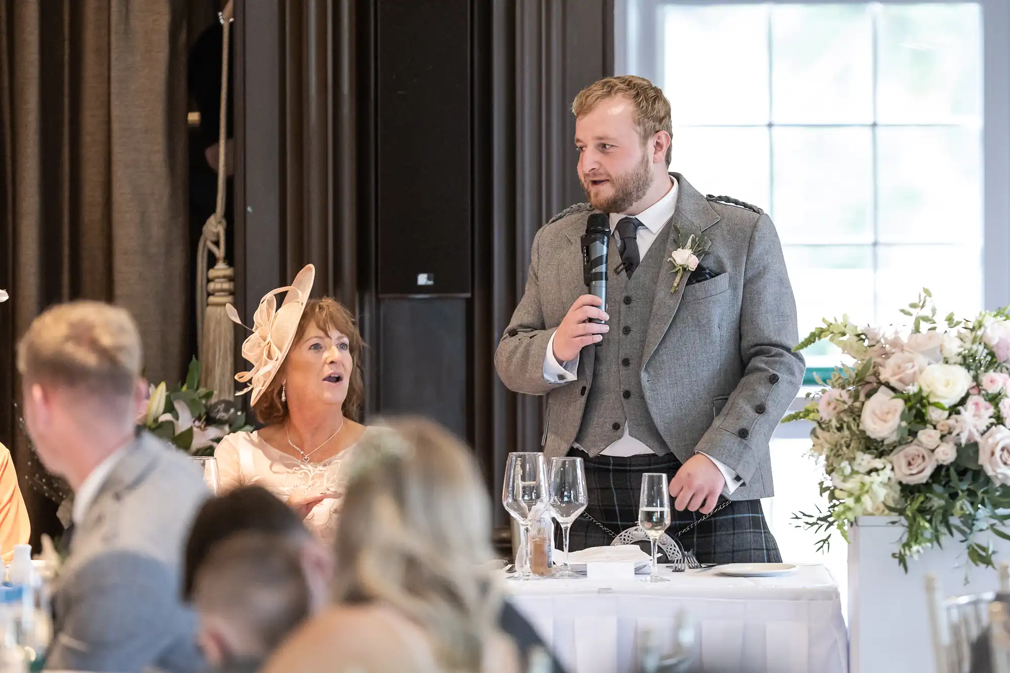 A man in a grey suit and kilt speaks into a microphone at a formal event, while a woman in a peach dress and matching hat sits beside him looking up. Guests are seated at tables.