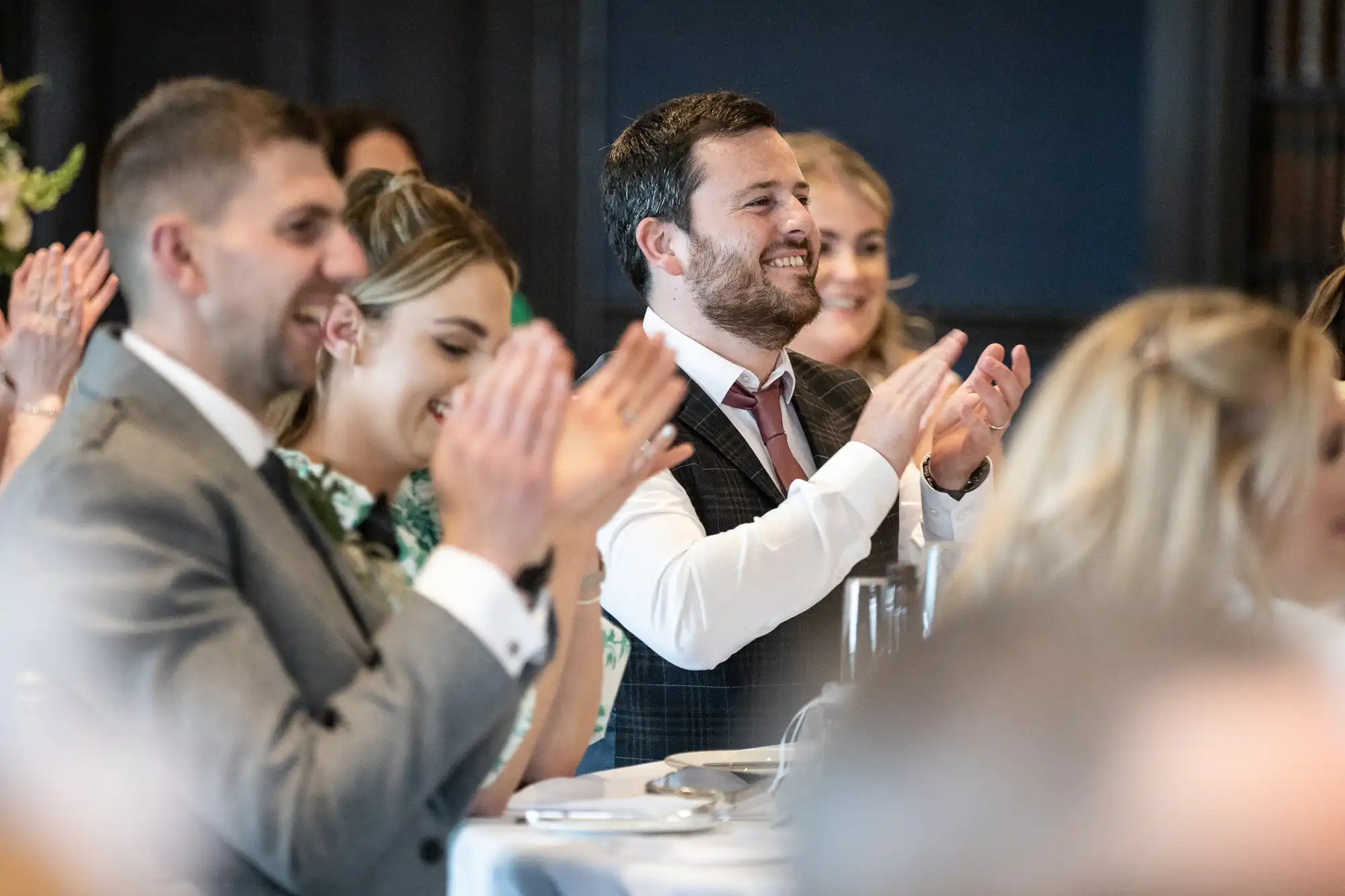 People seated at a table clapping and smiling, appearing to enjoy an event or performance. The setting suggests a formal or celebratory occasion.